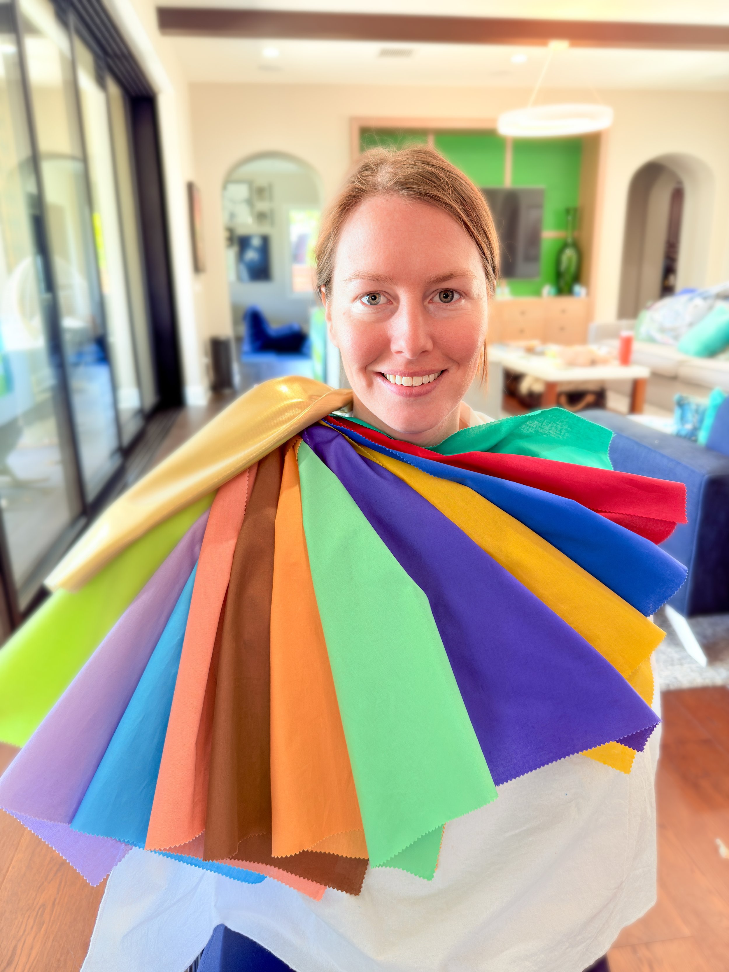 A person smiling with a rainbow-colored fabric color analysis drapes over their shoulders in a bright, modern room with large windows, a television, and decorative elements.
