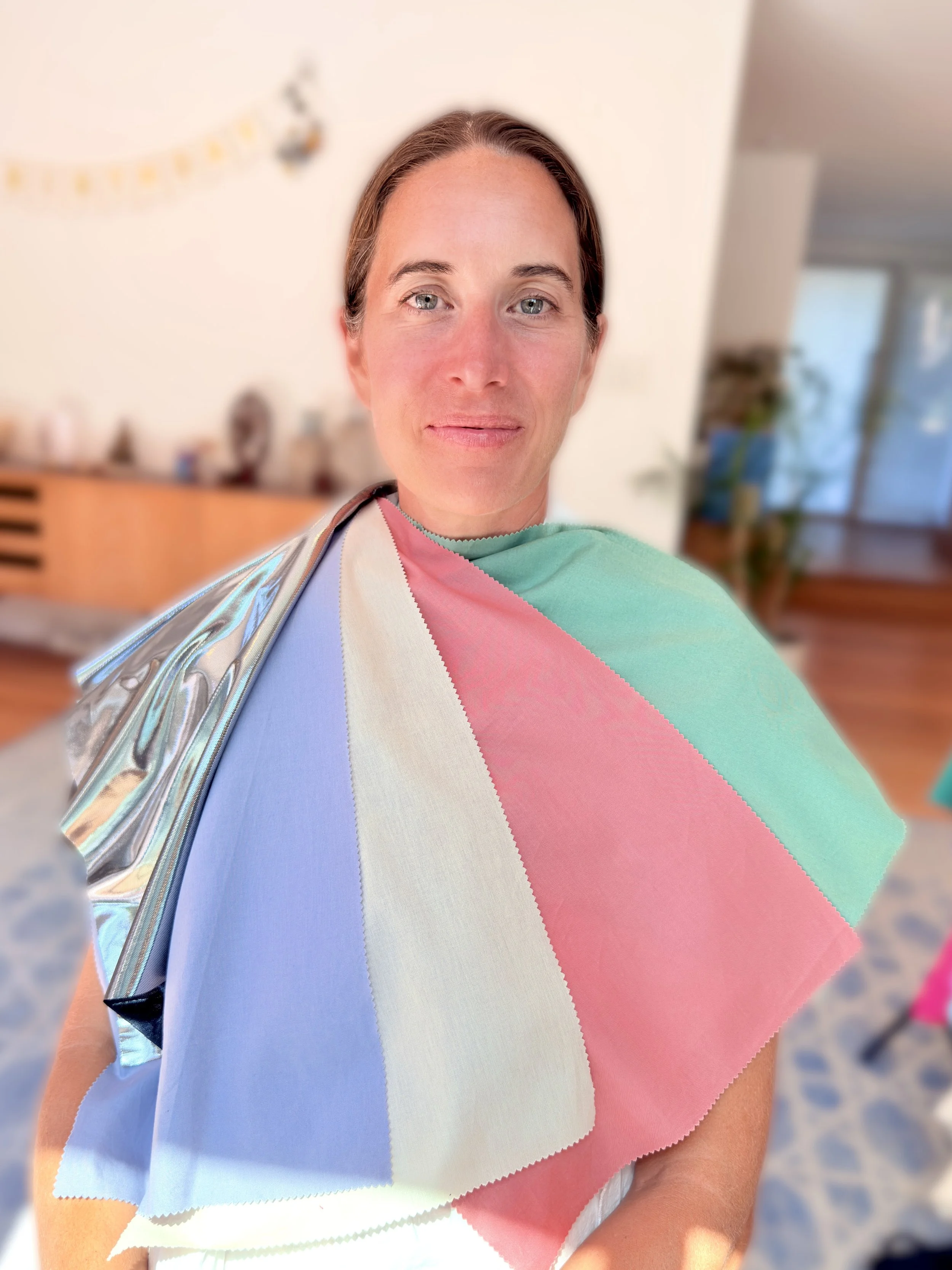 Close-up of a woman with blue eyes and brown hair wearing pastel-colored fabric swatches draped around her shoulders in a well-lit indoor space.