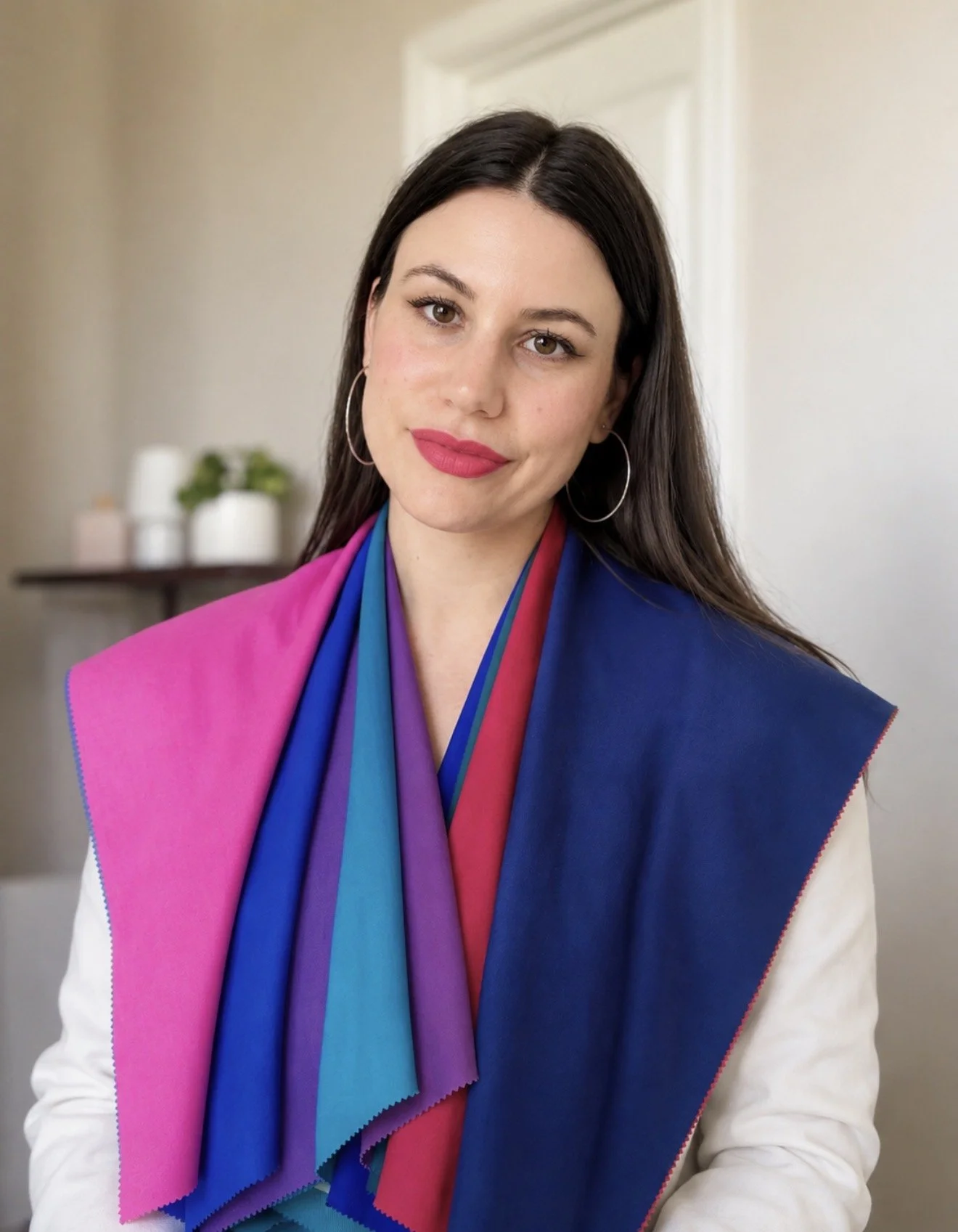 A woman with dark brown hair wearing a colorful color analysis drapes that includes pink, purple, blue, and red sections, and large silver hoop earrings, standing indoors with a neutral background featuring a table with white pots and green plants.