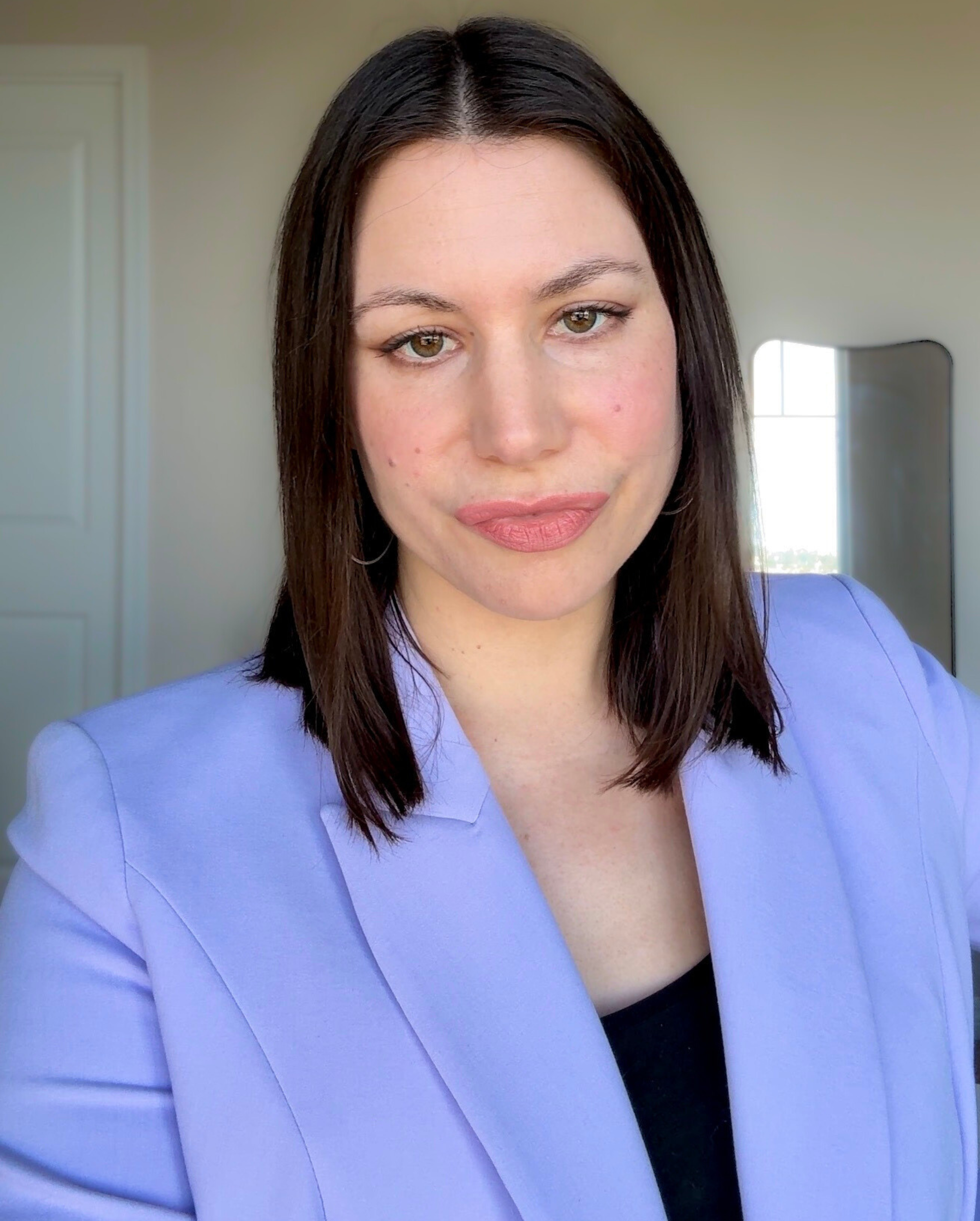 A woman with shoulder-length dark brown hair and fair skin, wearing a lavender blazer and black top, smiling softly in a well-lit room with a window and mirror in the background.