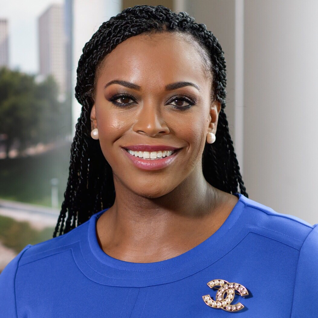 Professional woman wearing a blue dress with a double c/Chanel pin, smiling with dark pink lipstick, braided hair, against a light gray background.
