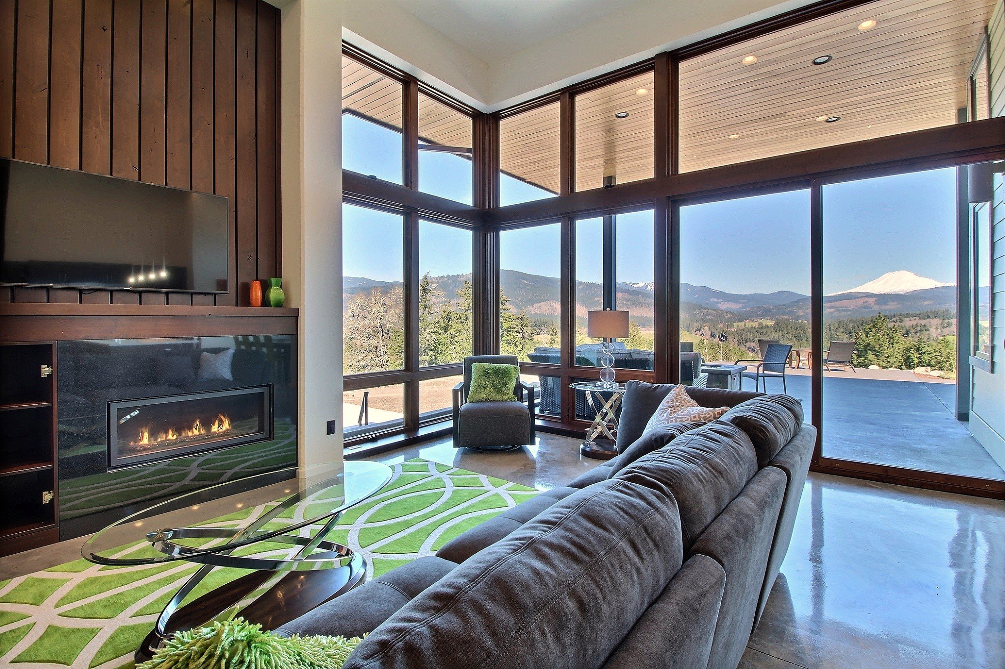 Modern living room with large windows, outdoor mountain view, sectional couch, glass coffee table, fireplace, and abstract rug.