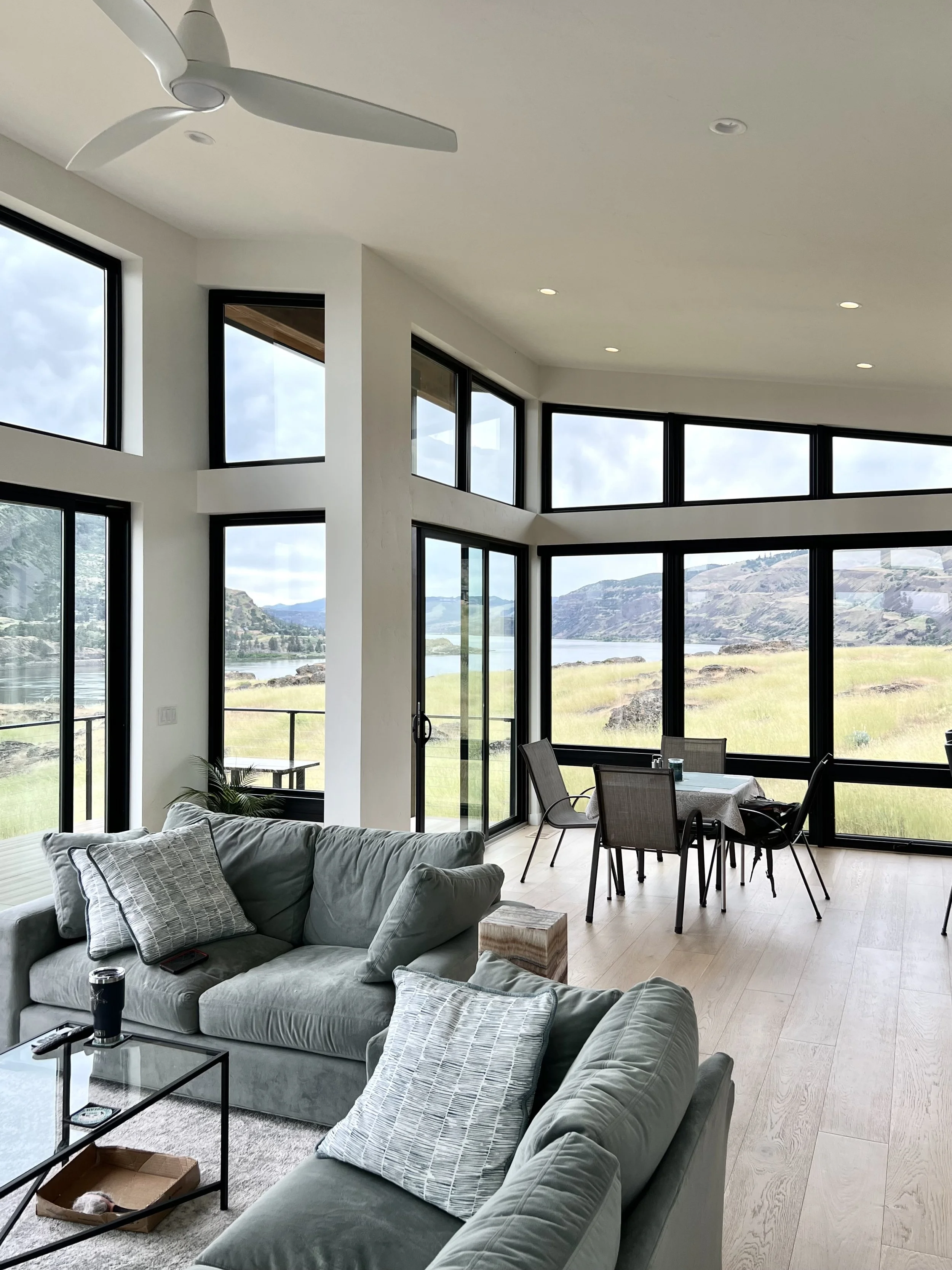 Modern living room with large windows, green sofas, glass coffee table, dining area, and ceiling fan, overlooking a natural landscape.