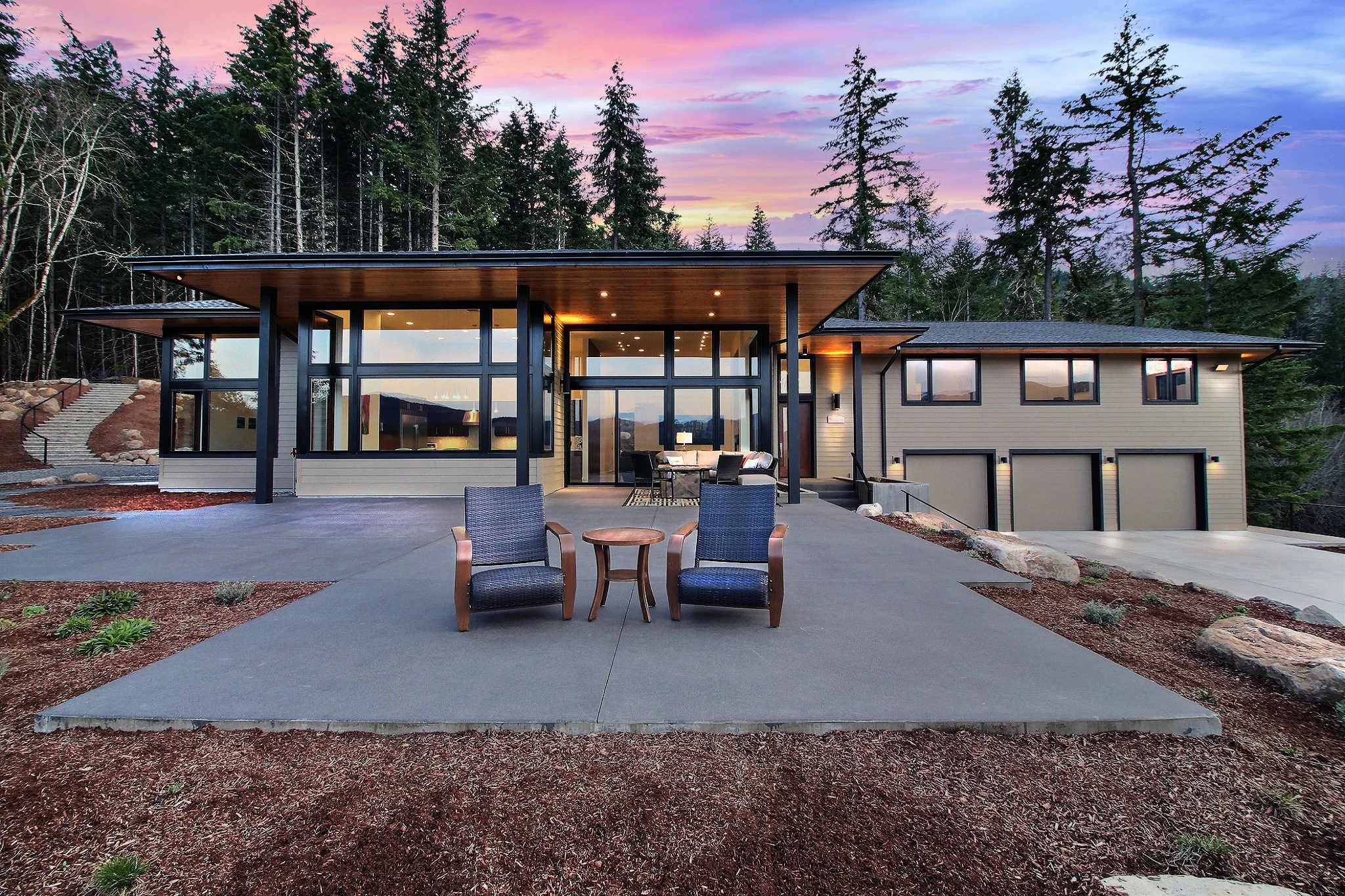 Modern house with large glass windows and patio, surrounded by trees, during sunset.