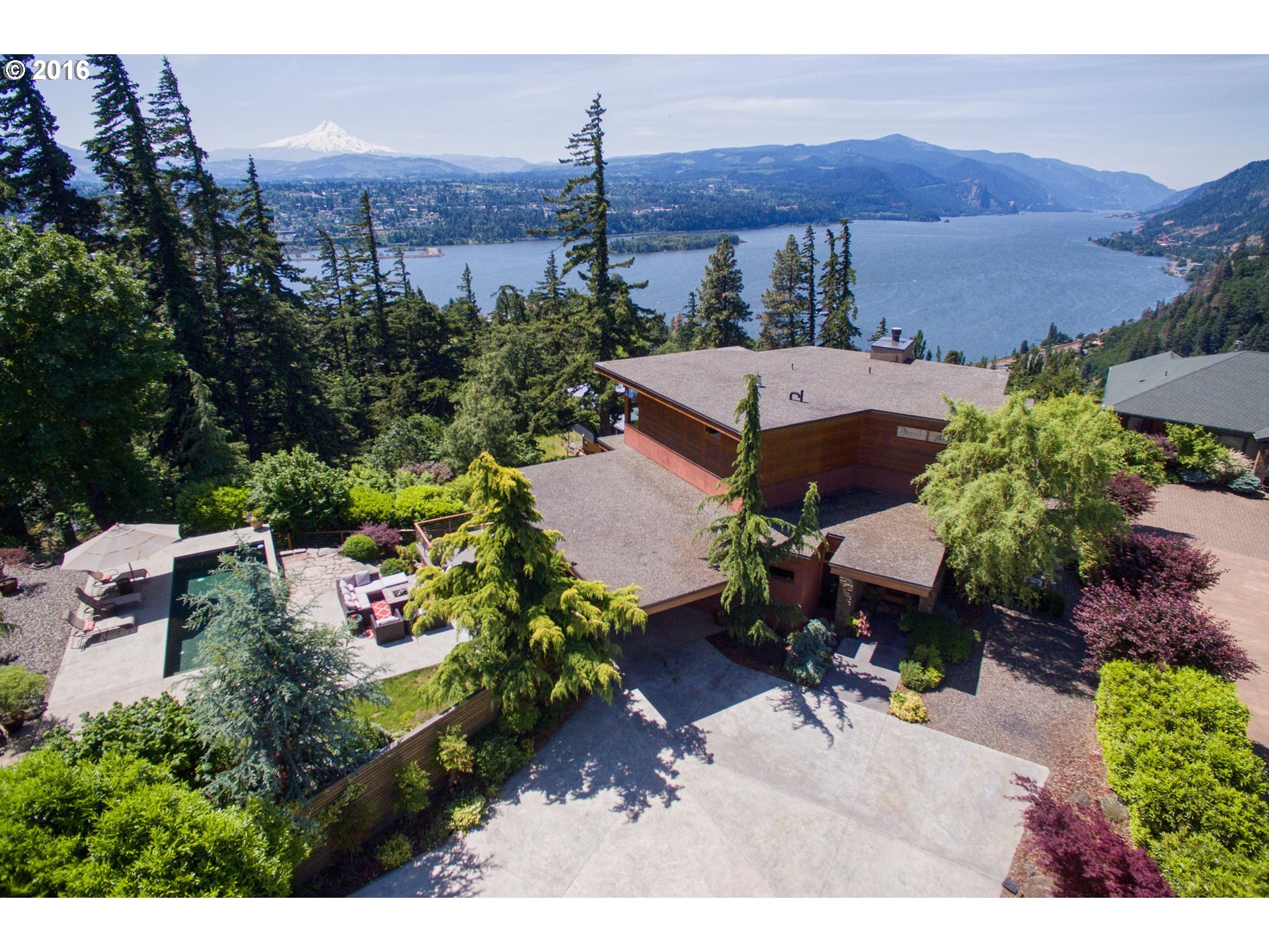 Aerial view of a modern house surrounded by lush greenery with a swimming pool, overlooking a scenic river and mountains.
