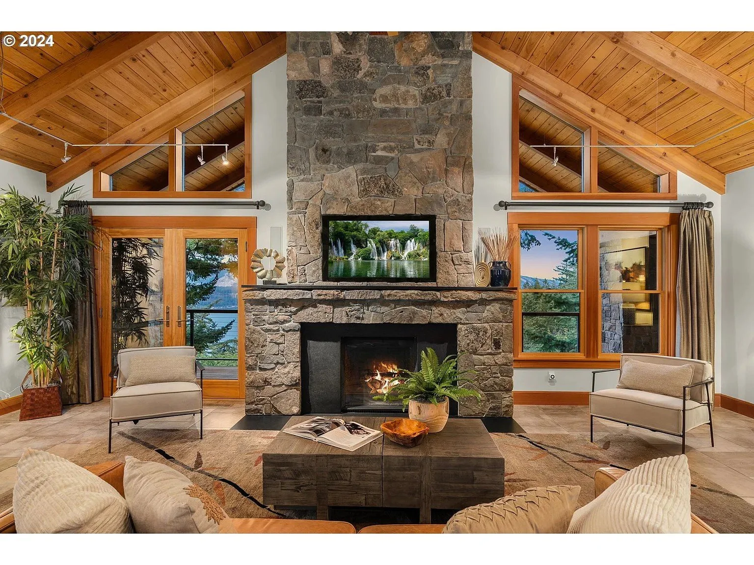 Cozy living room with high wooden ceiling, stone fireplace, two armchairs, a coffee table, plants, and large windows with outdoor views.
