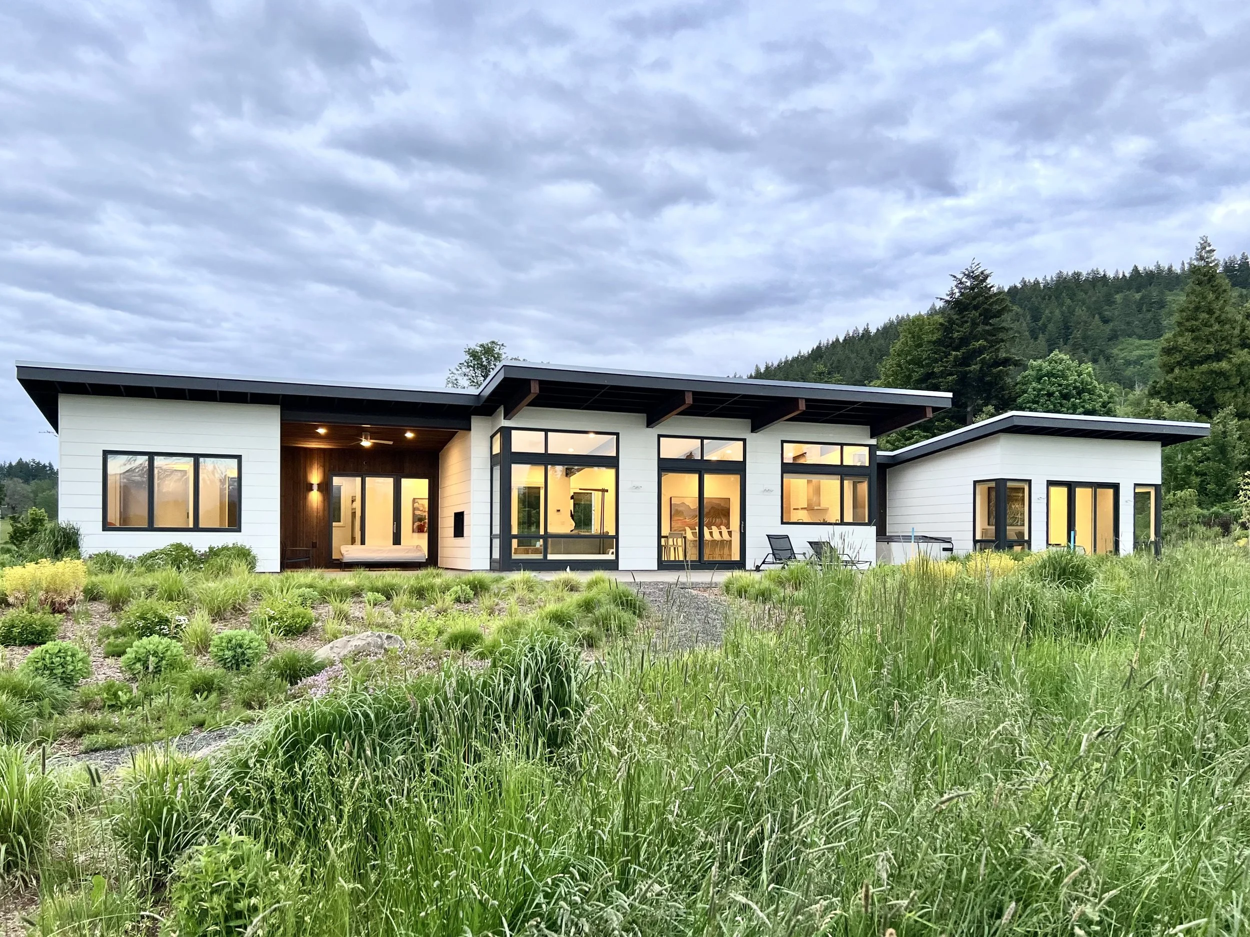 Modern single-story house with large windows, surrounded by grass and greenery, under a cloudy sky.