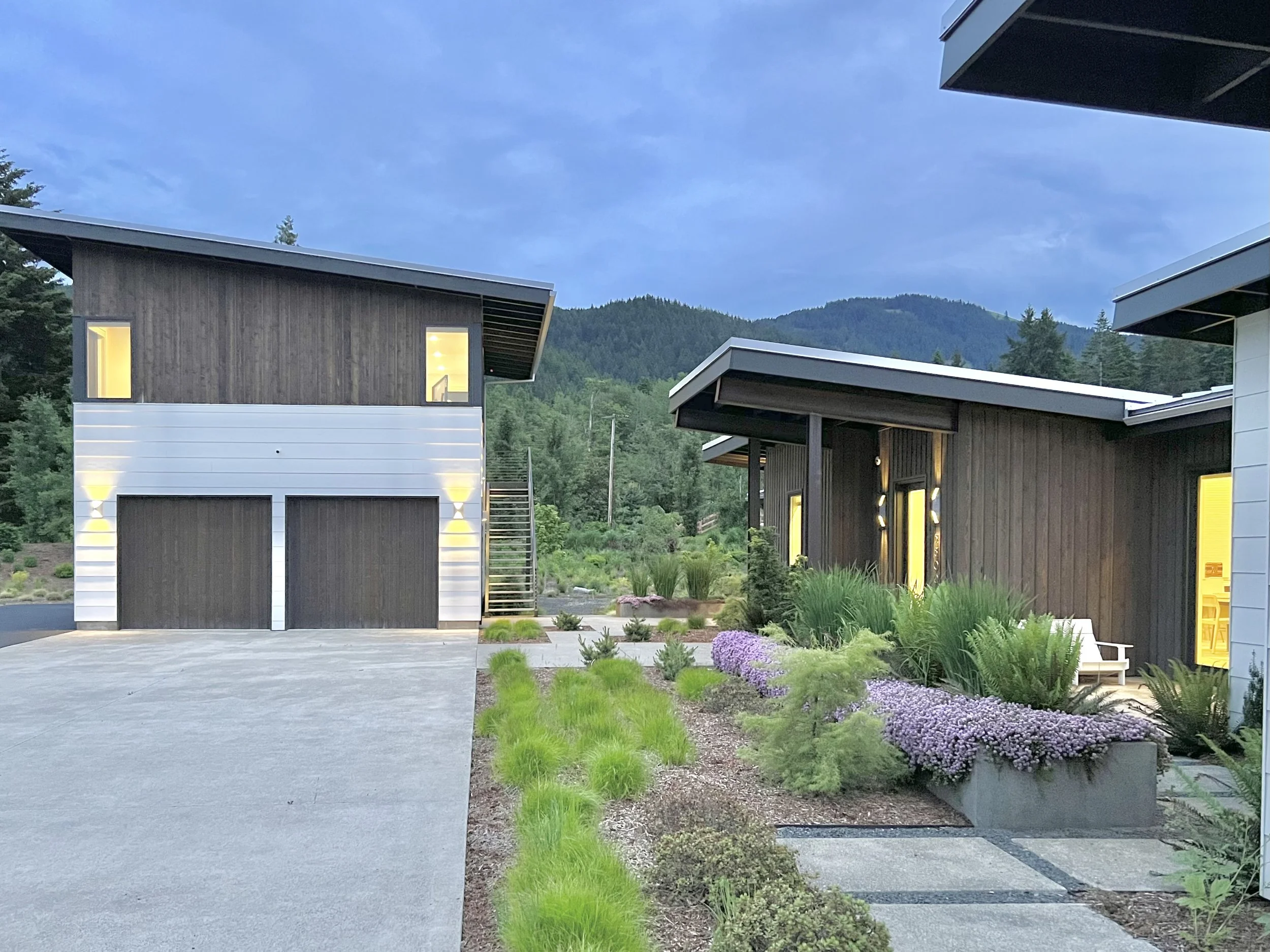 Modern house with wooden and metal exterior, surrounded by landscaped garden and mountain backdrop.
