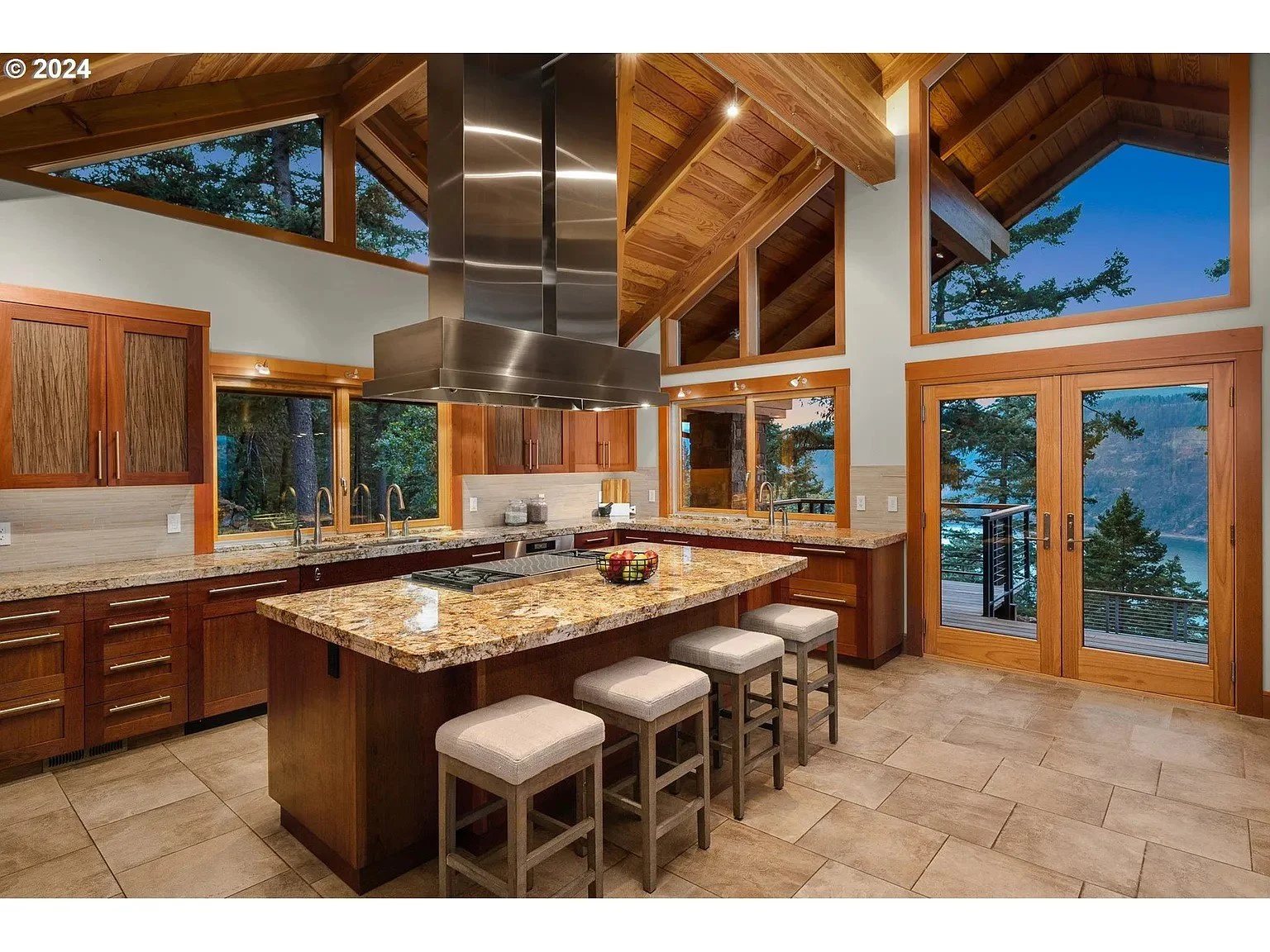 Modern kitchen with wooden cabinets, granite countertops, island with stools, large windows, stainless steel range hood, and view of trees.