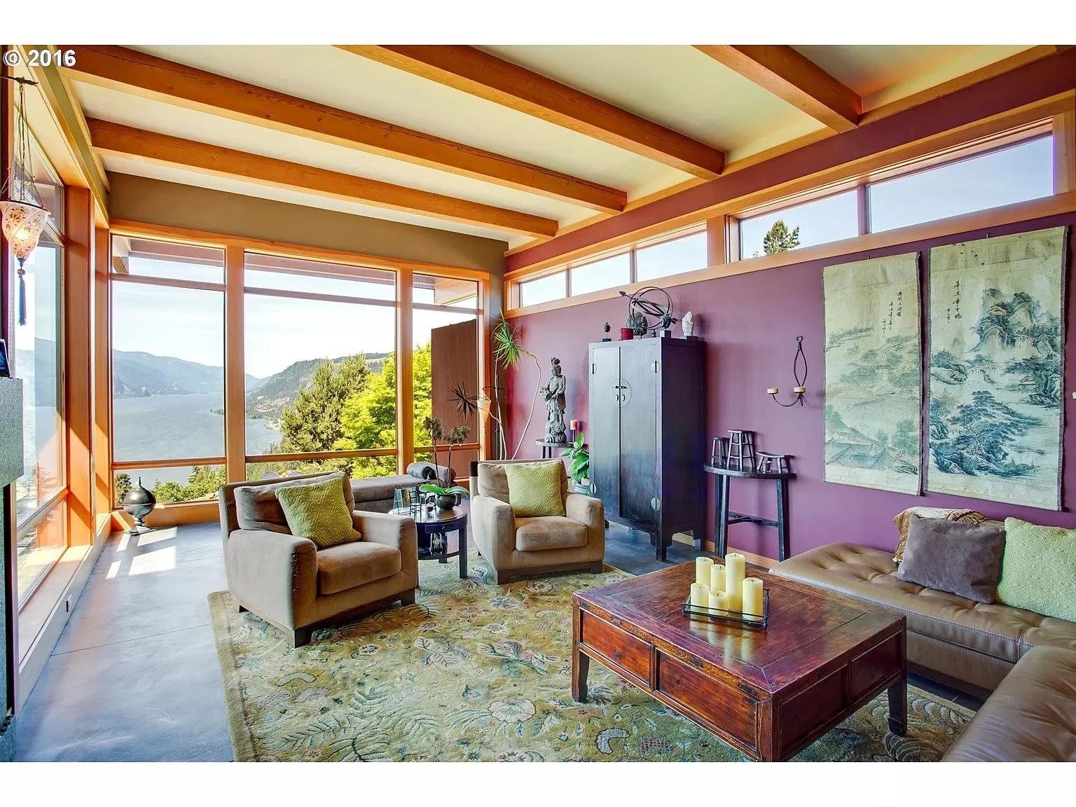 Modern living room with large windows overlooking a scenic view, including a lake and mountains. The room features brown furniture, two armchairs, a wooden coffee table with candles, an Asian-style cabinet, decorative wall art, and an indoor plant.