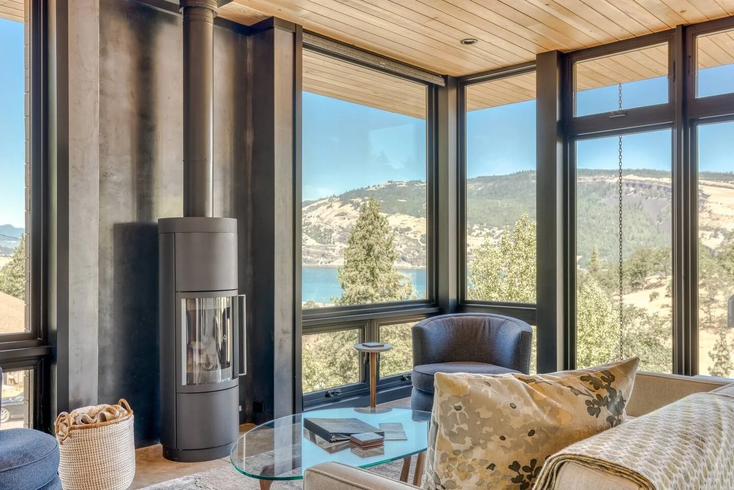 Modern living room with panoramic windows, a black wood-burning stove, blue armchair, glass coffee table, and scenic nature view.