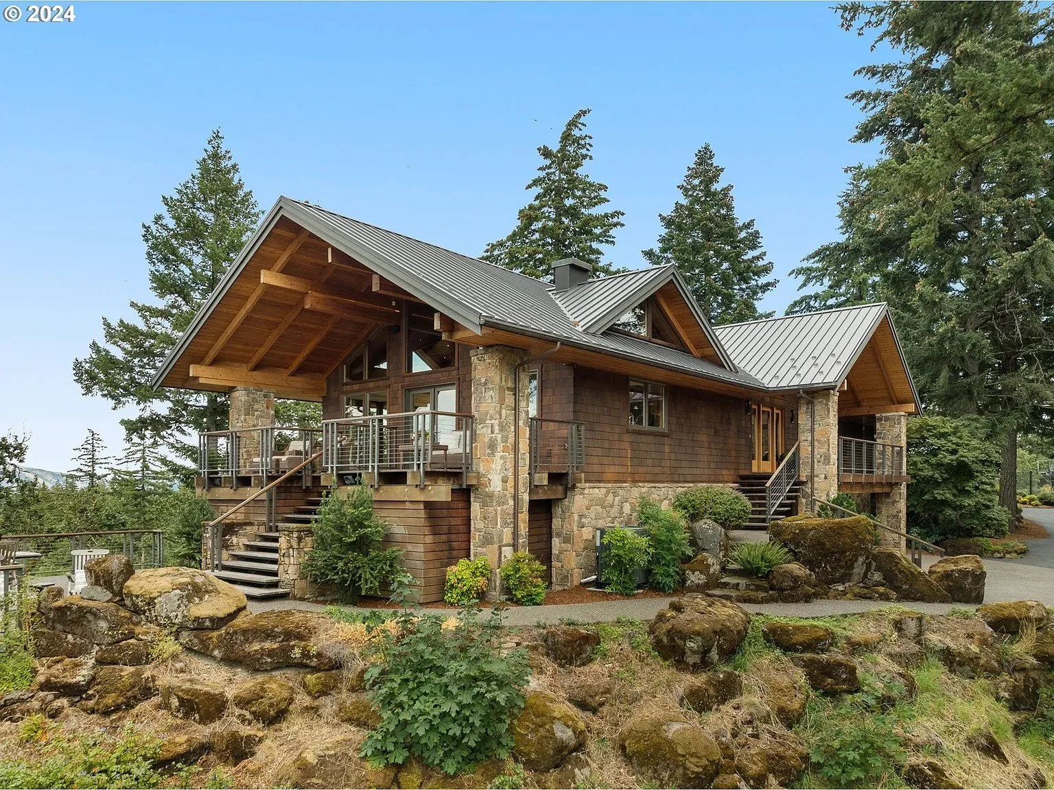 A modern rustic house with a wooden and stone exterior, surrounded by trees and landscaping, with a metal roof and a deck.