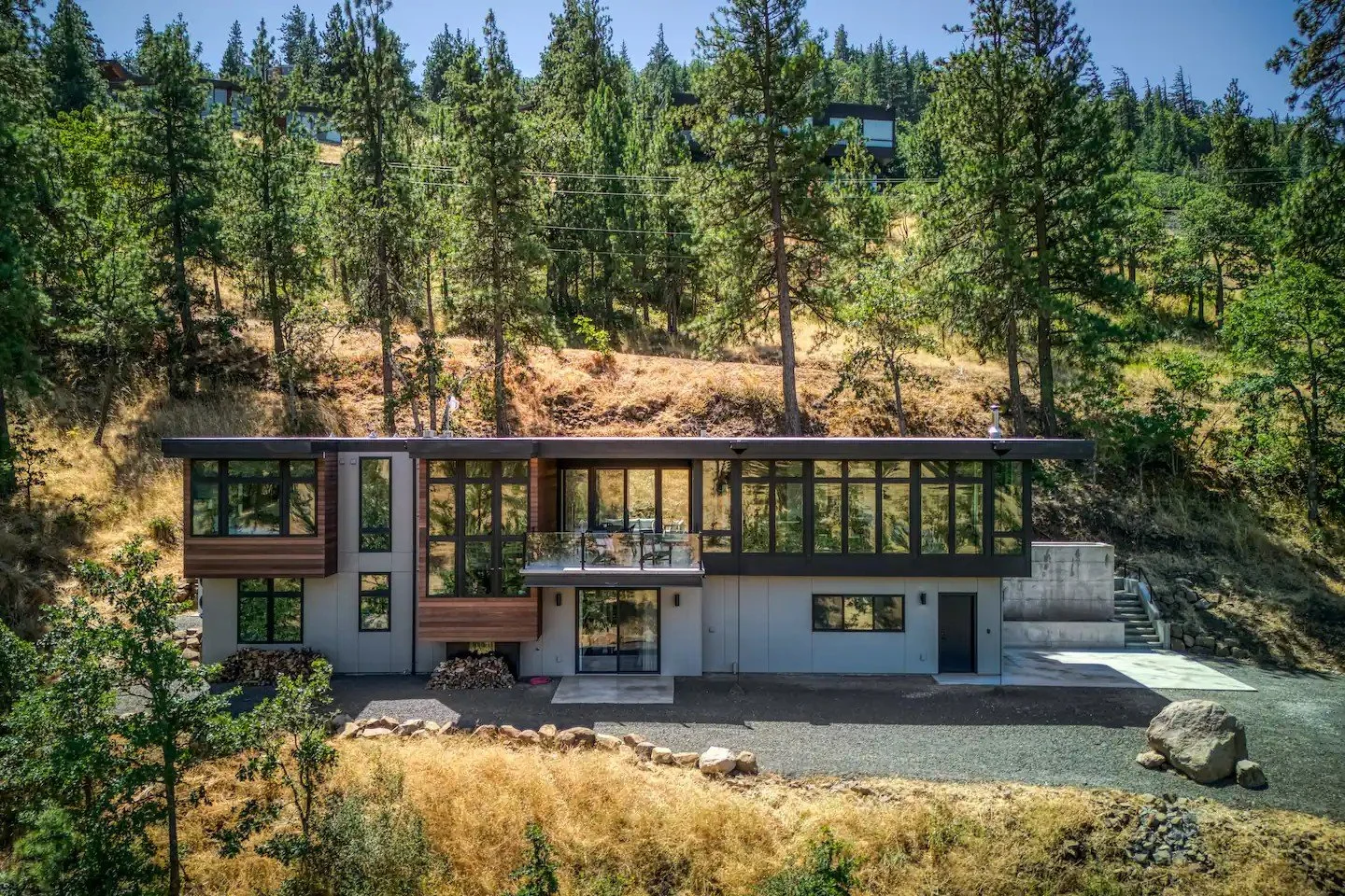 Modern two-story house with large windows surrounded by forest and hillside.