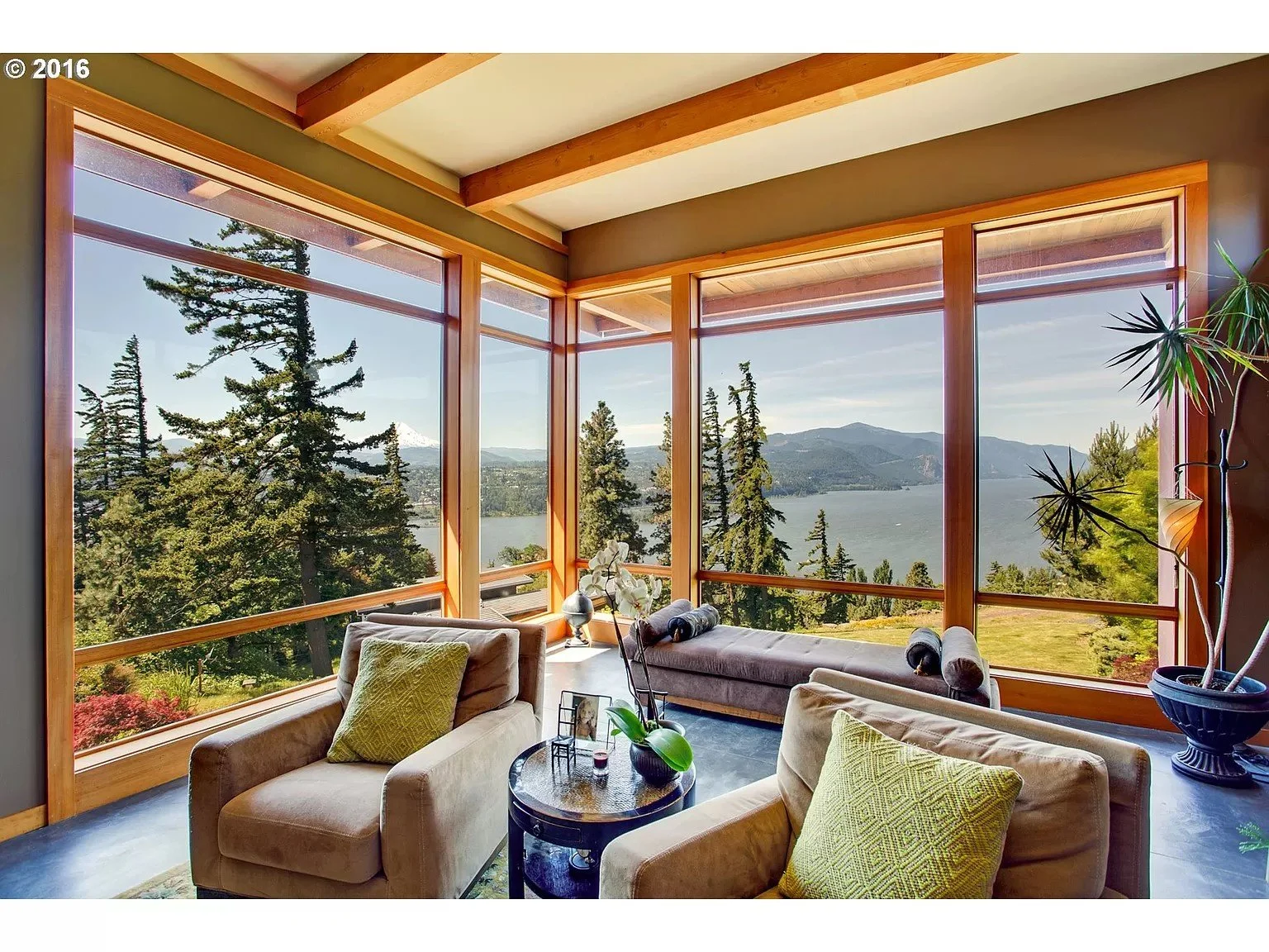 Modern living room with large windows, offering a panoramic view of a river, trees, and distant mountains. Interior includes beige armchairs, a small round table, and green plants.