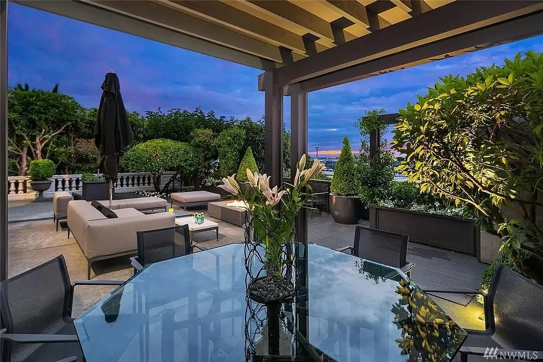 Outdoor patio with glass table, cushioned seating, potted plants, under a pergola at sunset.