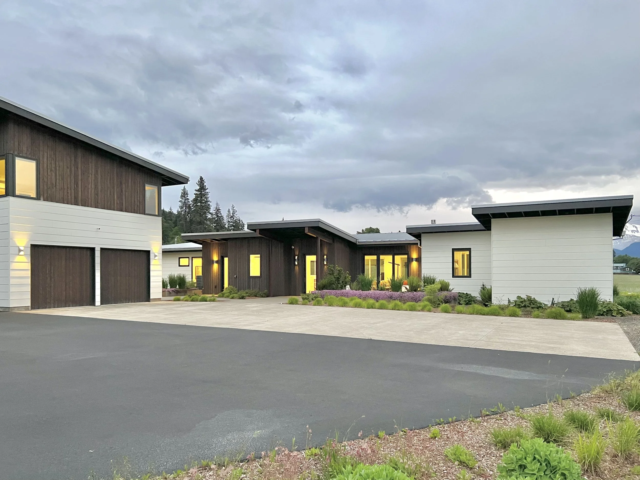 Modern house exterior with wood and white paneling, large windows, manicured landscaping, and overcast sky.