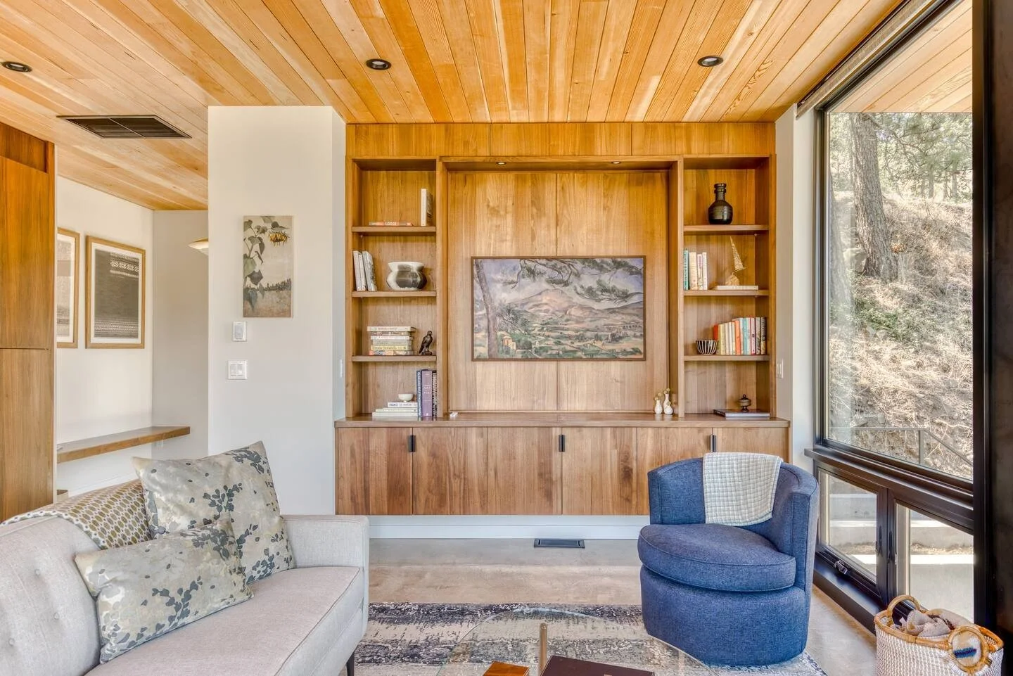 Modern living room with wood ceiling, built-in bookshelves, a sofa with decorative pillows, round blue chair, large window, and wall art.
