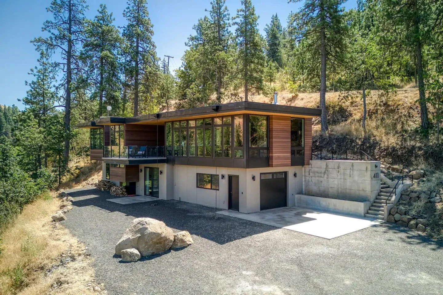 Modern two-story house with large glass windows in a forest setting, surrounded by tall pine trees; features include a gravel driveway, concrete steps, and wooden exterior accents.