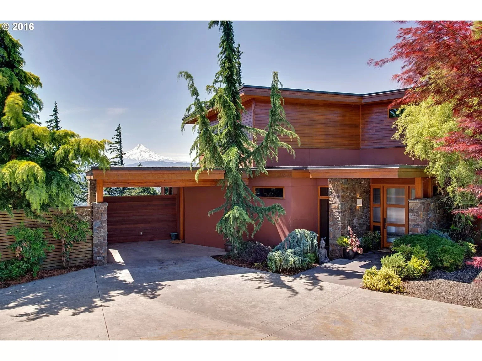 Modern house with wood and stone facade, surrounded by lush greenery, and a snow-capped mountain visible in the background.