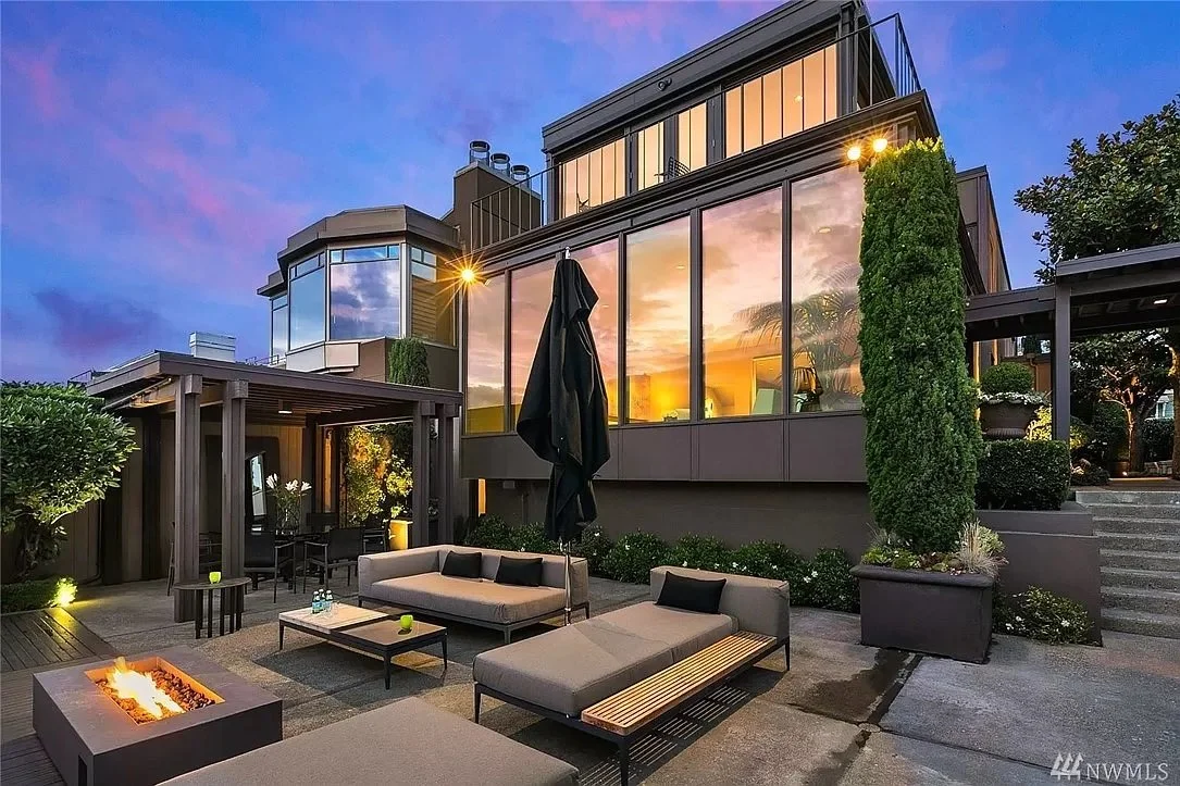 Modern house exterior with large windows, outdoor seating area, fire pit, and evening lighting.