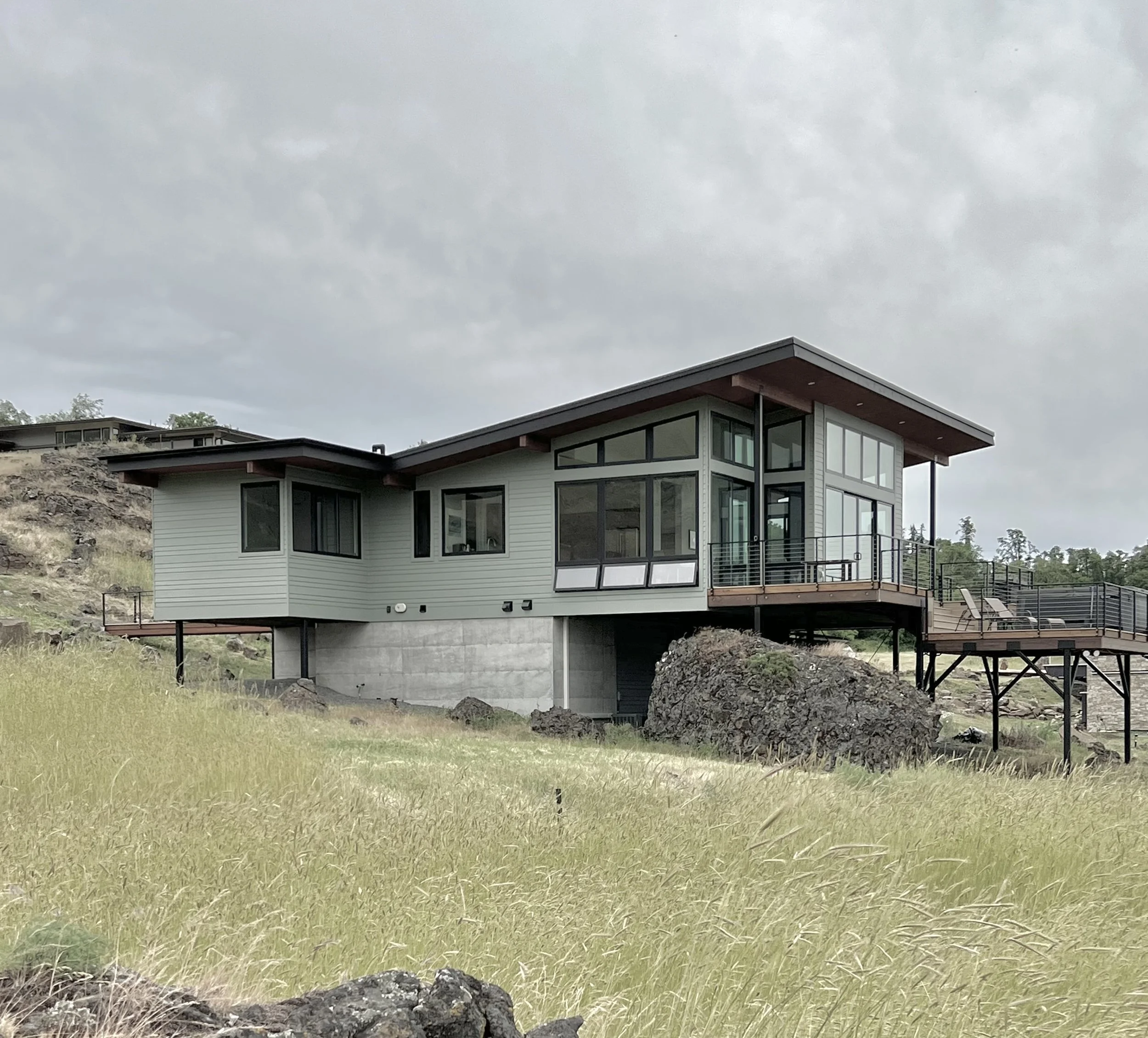 Modern hillside house with large windows and elevated deck, surrounded by grass and rocks.