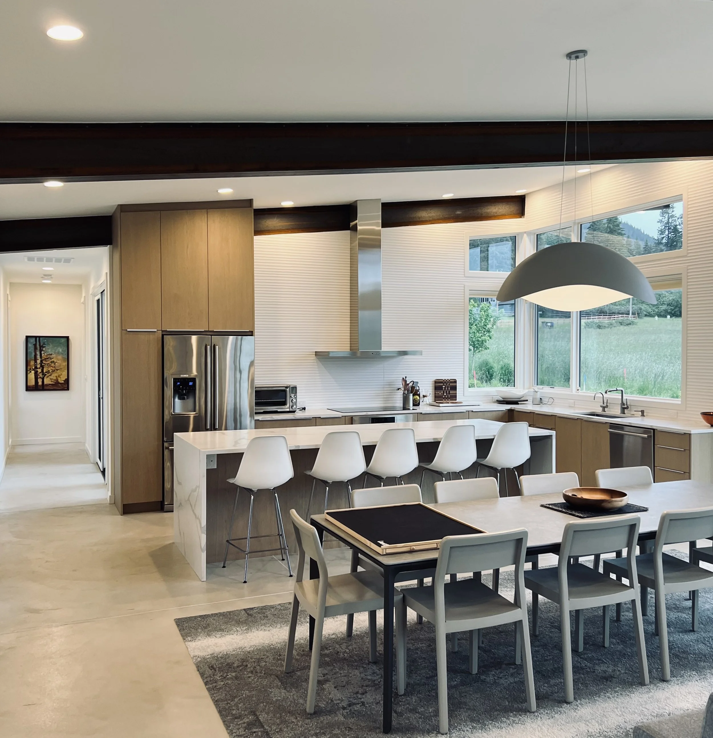 Modern kitchen and dining room with wooden cabinets, stainless steel appliances, a large dining table with chairs, and a central island with bar stools. A large window offers a view of greenery outside and a contemporary pendant light hangs above the table.