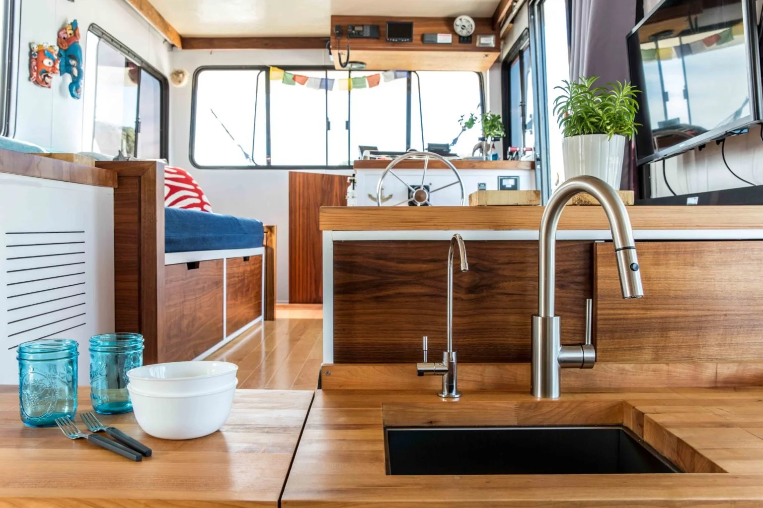 Interior of a houseboat featuring wooden cabinetry, a kitchen sink with a modern faucet, and a living area with blue cushions. There are plants, colorful prayer flags, and a steering wheel visible in the background.