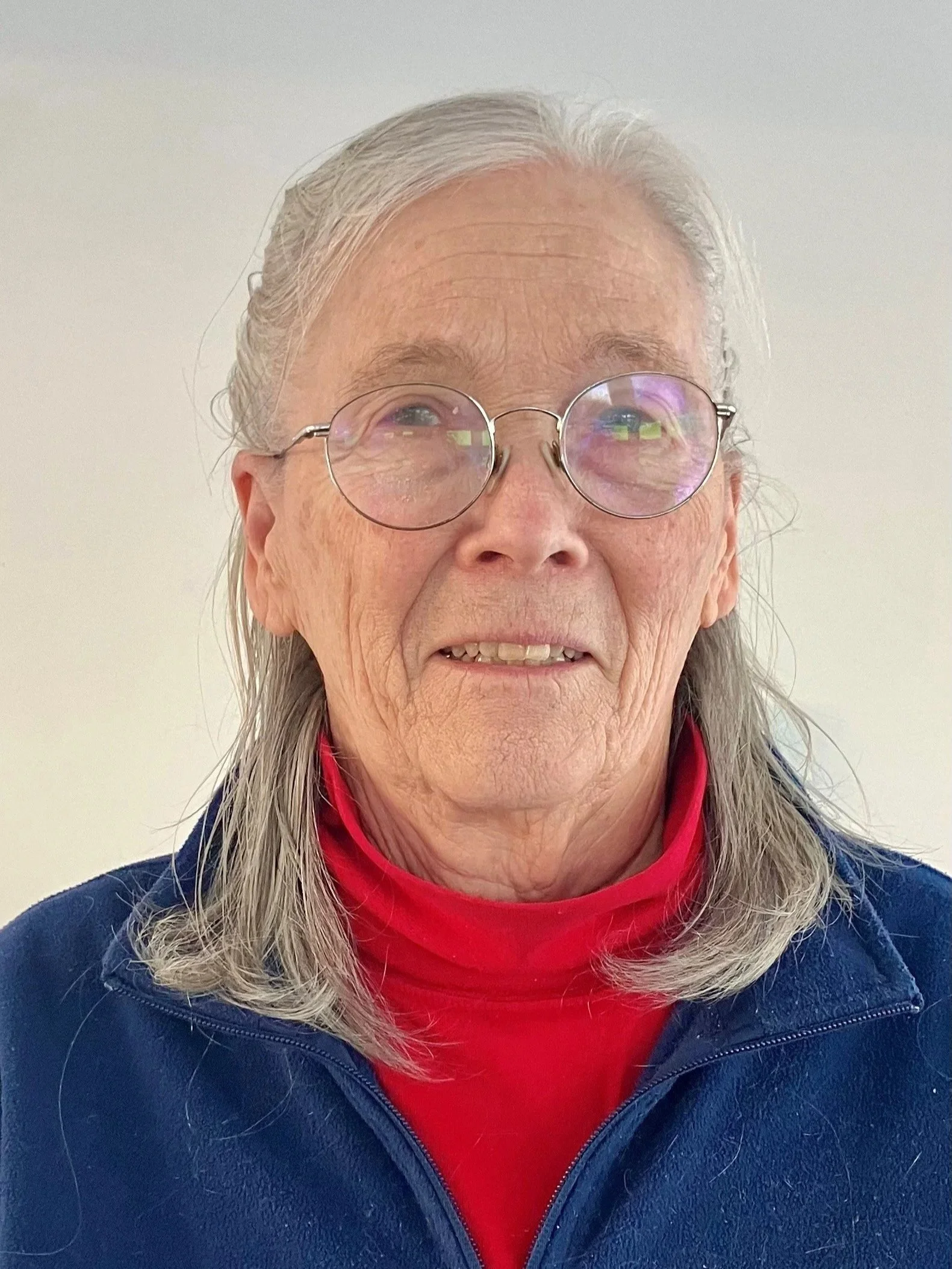 Close-up of an elderly woman with long gray hair, wearing glasses, smiling, and showing some facial wrinkles.
