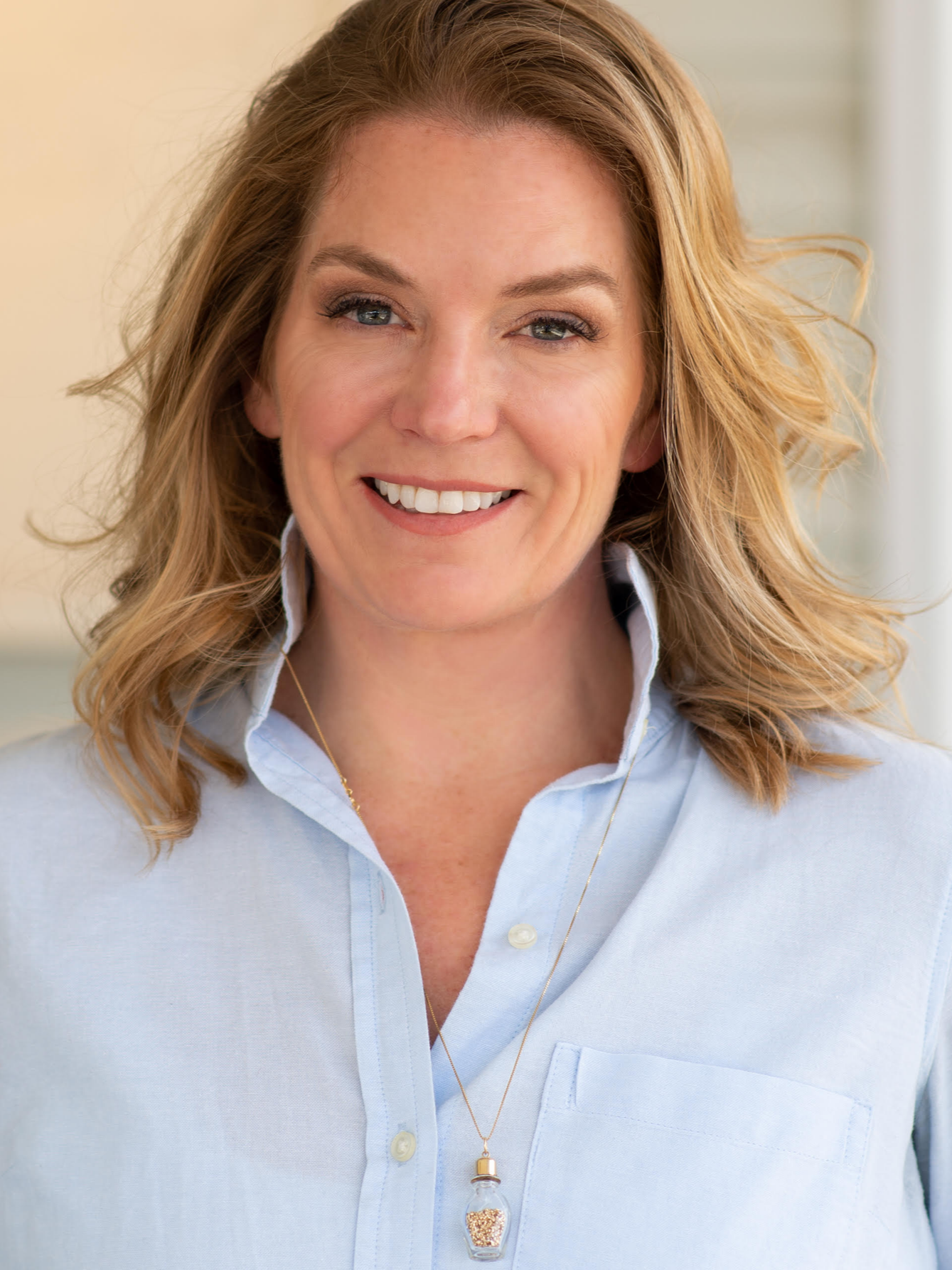 A woman with long, wavy hair smiling outdoors in a park on a sunny day, wearing a white shirt.