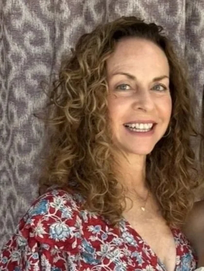 A woman with long curly hair wearing sunglasses and a dark blue sleeveless top standing outside near a railing with trees and a parking lot in the background.