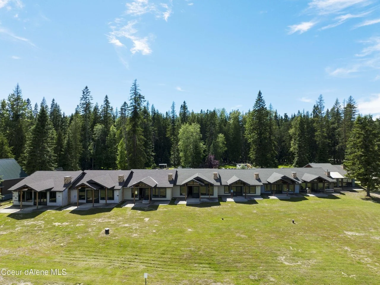 A row of modern single-story homes with brown roofs and large windows, set on a grassy field with a backdrop of tall pine trees under a clear blue sky.