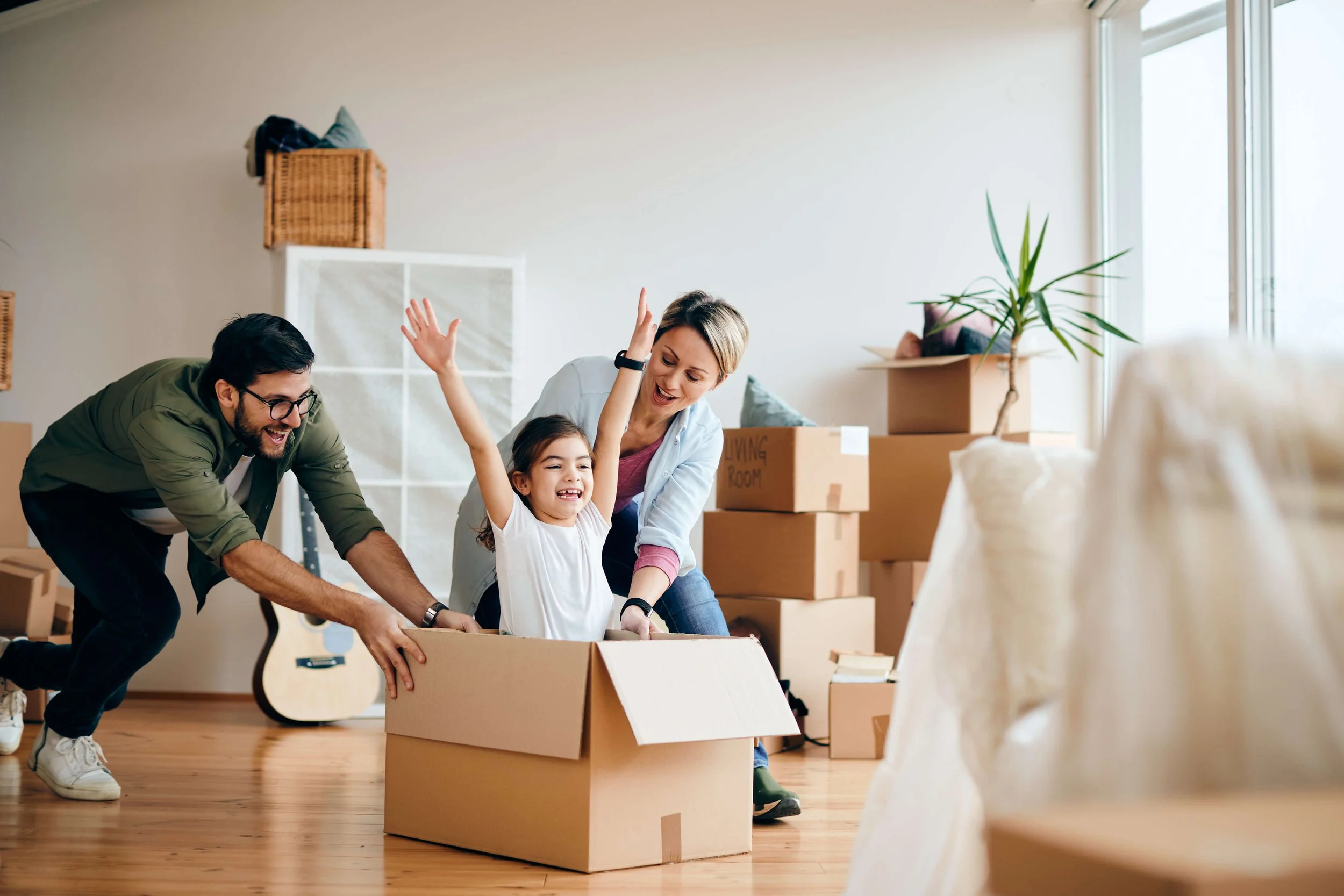 A family of three, a man, a woman, and a young girl, having fun as they push a girl inside a cardboard box during a move in a brightly lit room with moving boxes and a guitar in the background.