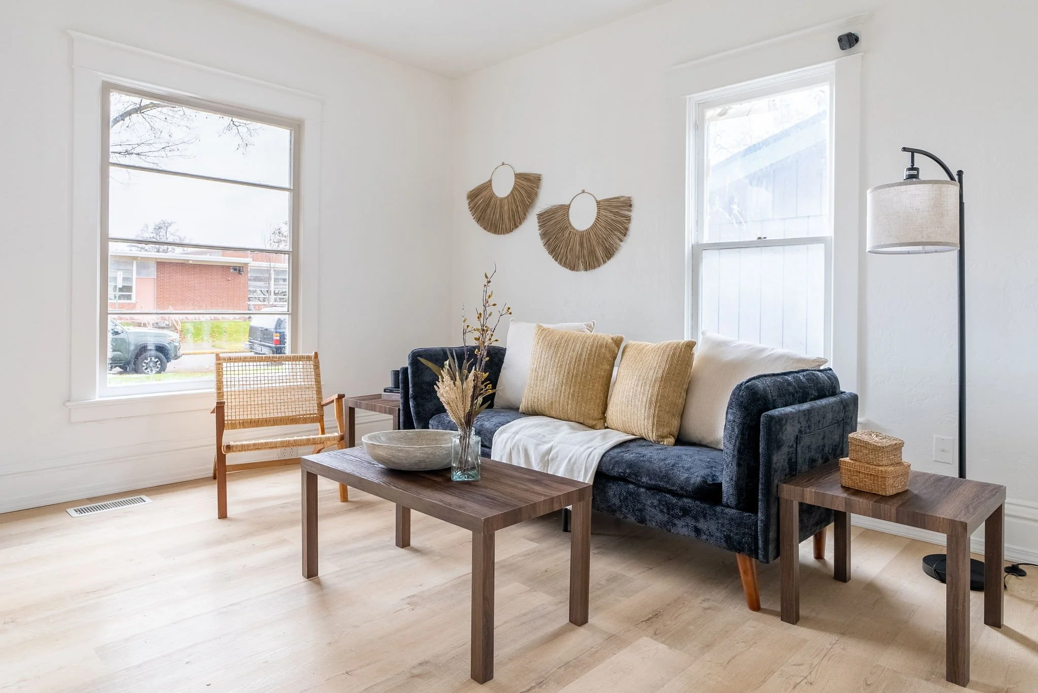 Living room with white walls, two large windows, and light wood flooring. Contains a dark blue sofa with beige and cream pillows, a wooden coffee table with a glass vase and dried flowers, a small wooden side table with woven baskets, a floor lamp with a beige lampshade, and wall art made of circular straw and mirror decorations.