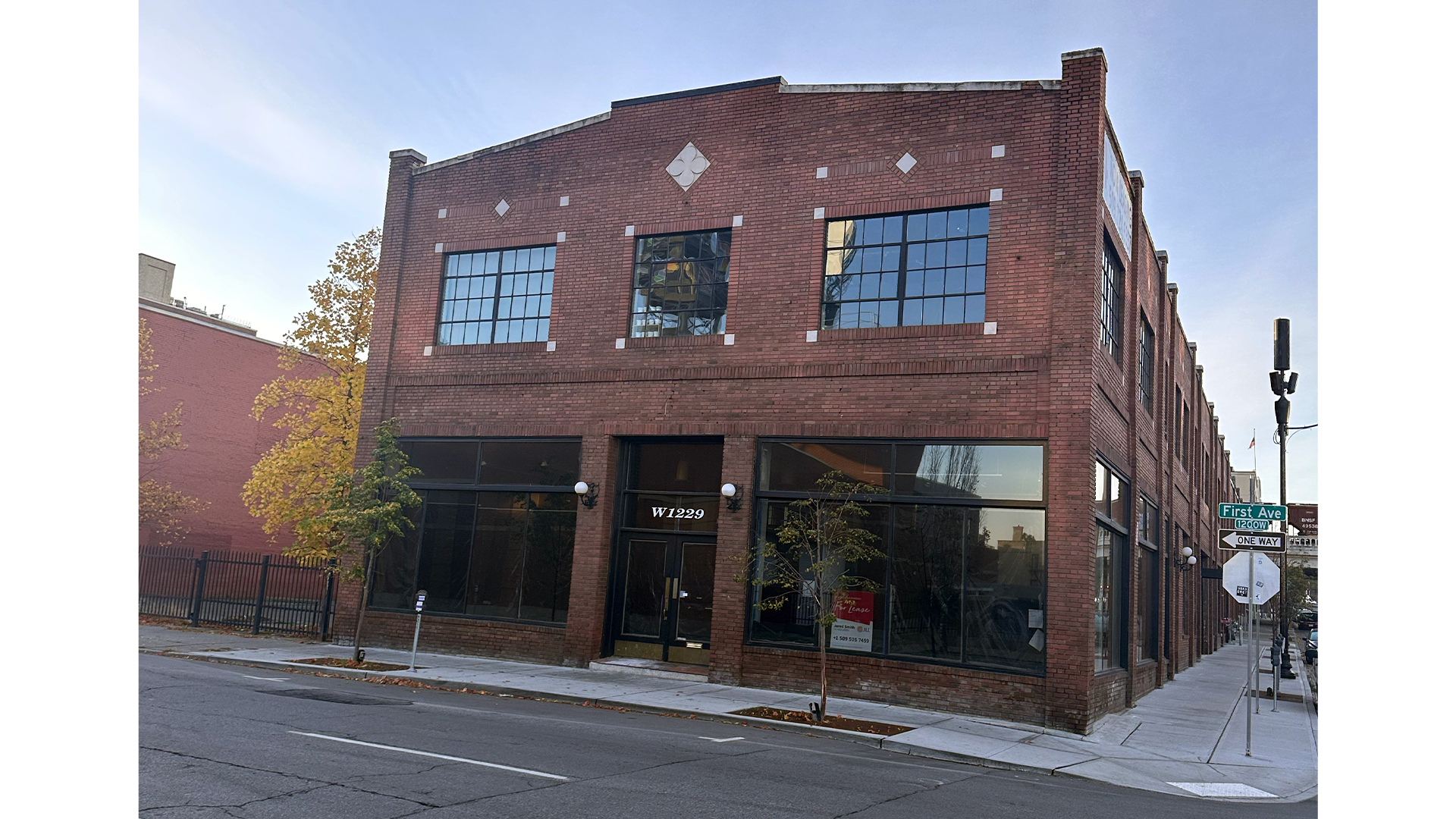 A three-story brick building on a city street corner with large ground-floor windows and the number W1229 above the door. Street signs indicate First Ave and a one-way street.