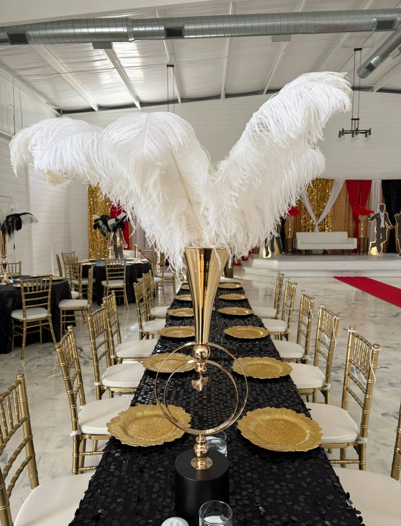 Event table setting with gold and black theme, featuring a centerpiece of white ostrich feathers in a gold vase surrounded by gold chairs and plates.