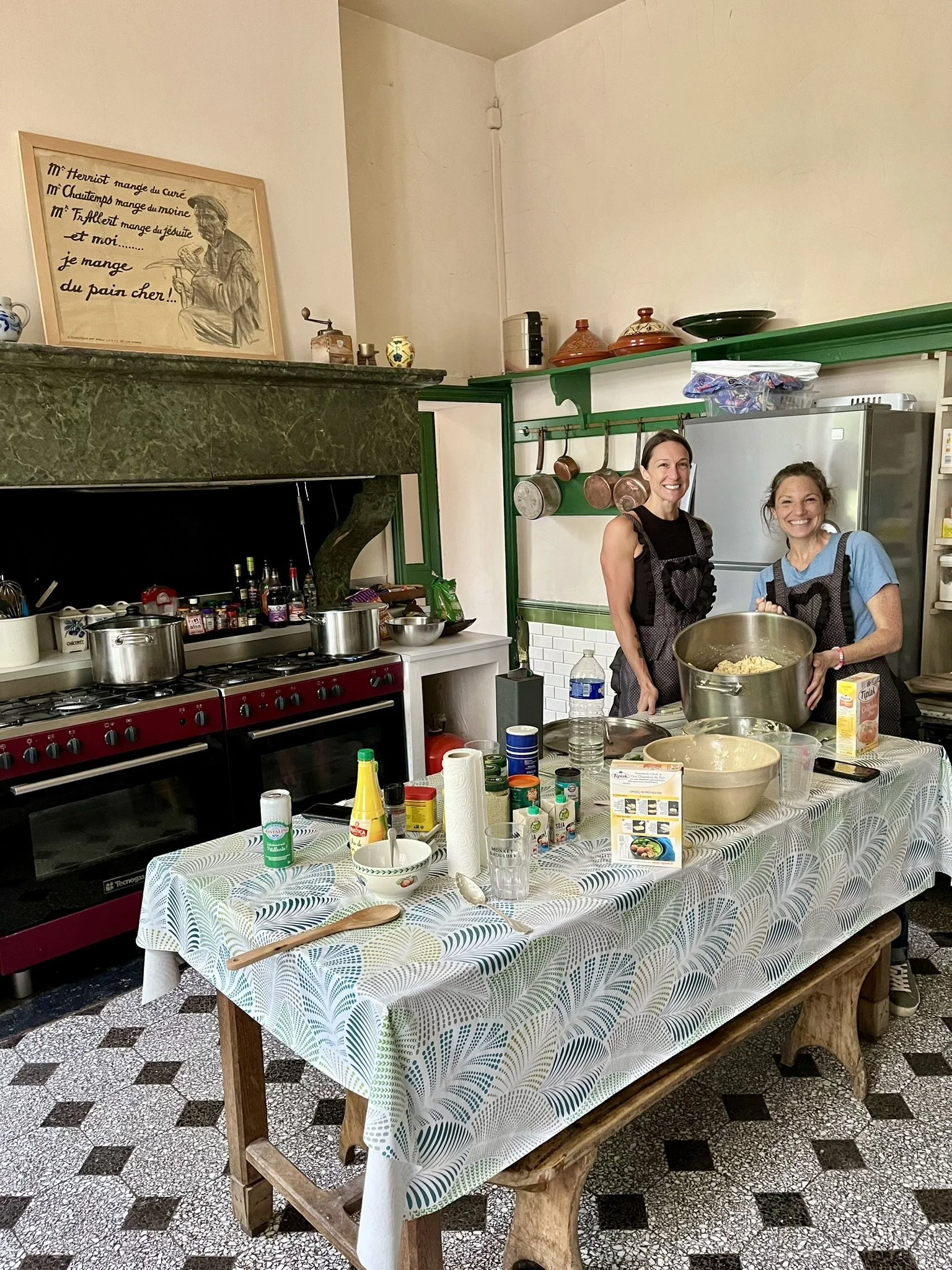 Two women in an apron cooking in a kitchen, standing behind a table filled with ingredients and kitchen tools, smiling at the camera.