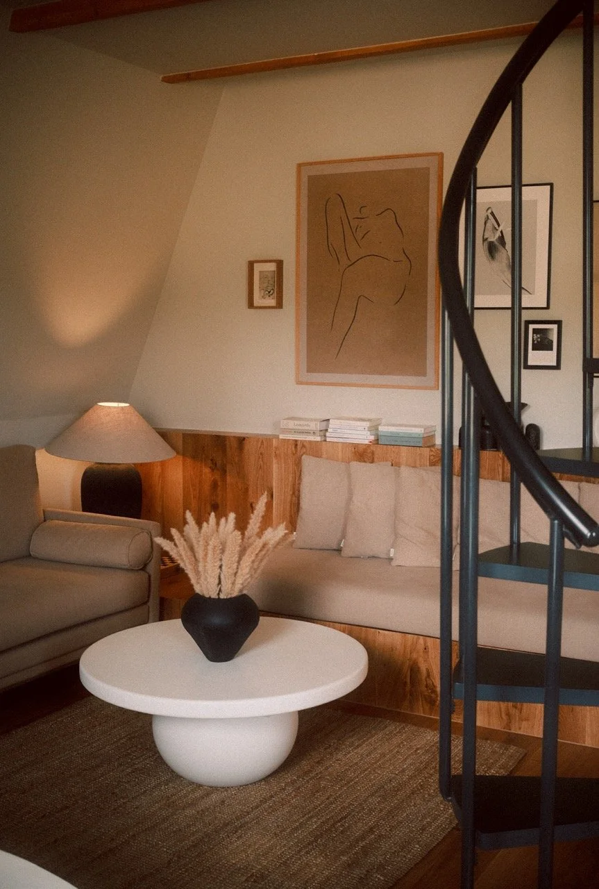Contemporary living room with beige sofas, a black vase with pampas grass on a round white coffee table, a wooden wall panel, framed line art and photographs on the wall, a table lamp, and a spiral staircase.