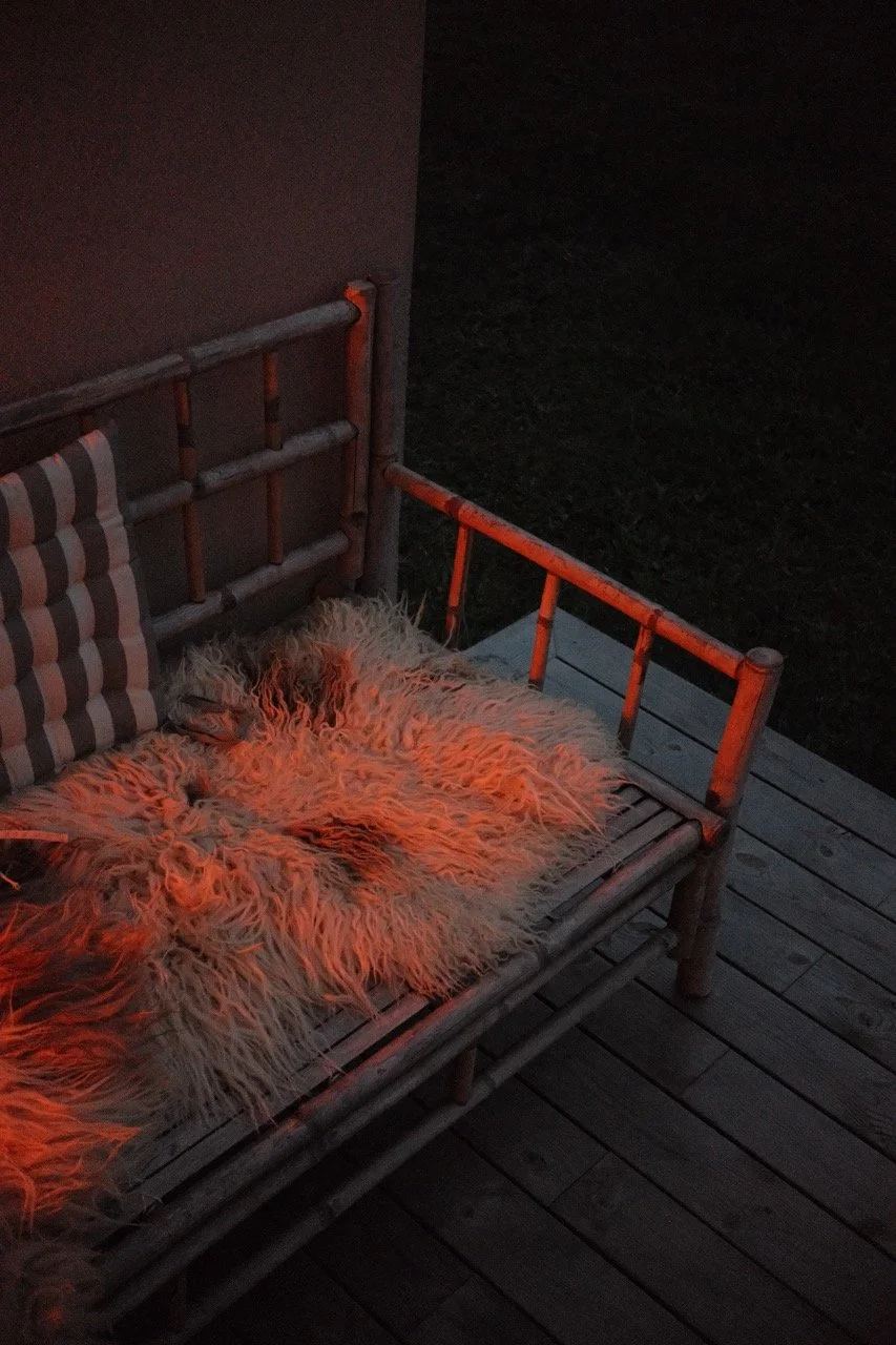 A wooden outdoor bench with striped cushions and a fluffy sheepskin rug in a dimly lit setting, illuminated with a warm reddish glow.