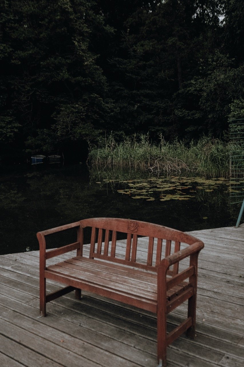 A wooden bench on a deck by a pond with lily pads, surrounded by trees at dusk or night.