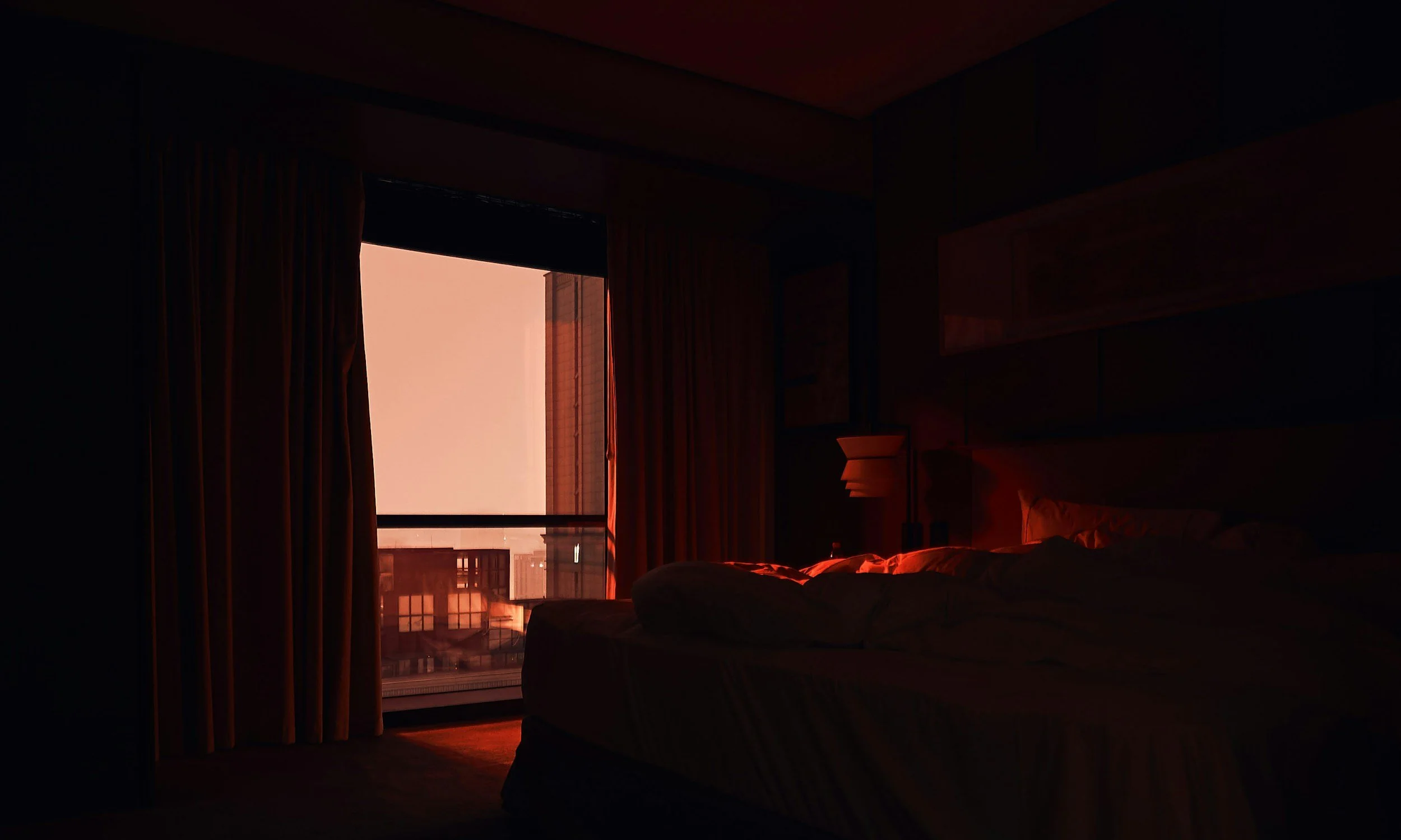Dark bedroom with a partially open window showing a sunset sky and cityscape outside, bed unmade, curtains drawn.