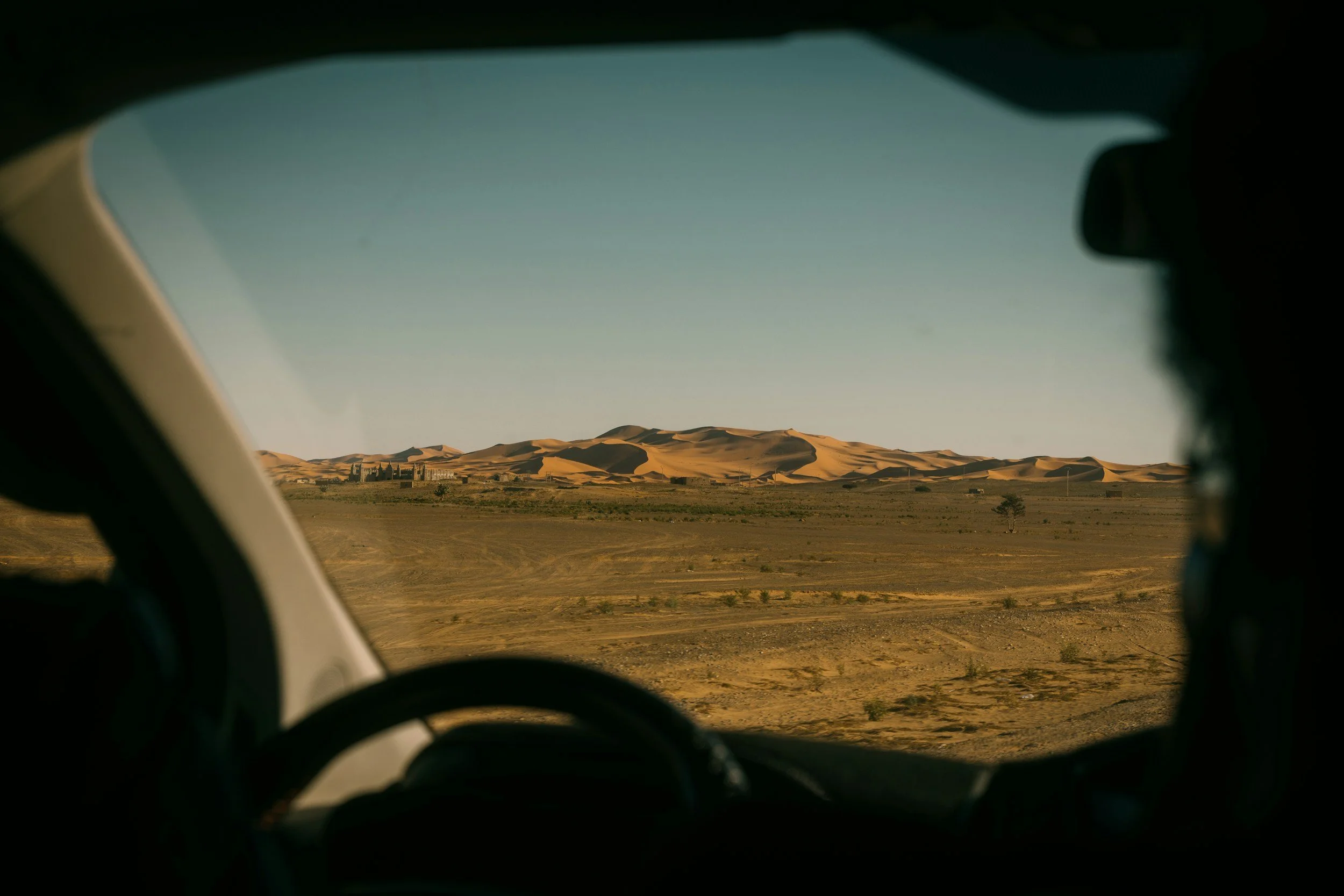 View of sand dunes in a desert landscape through the windshield of a vehicle, with the interior silhouette of the driver visible.