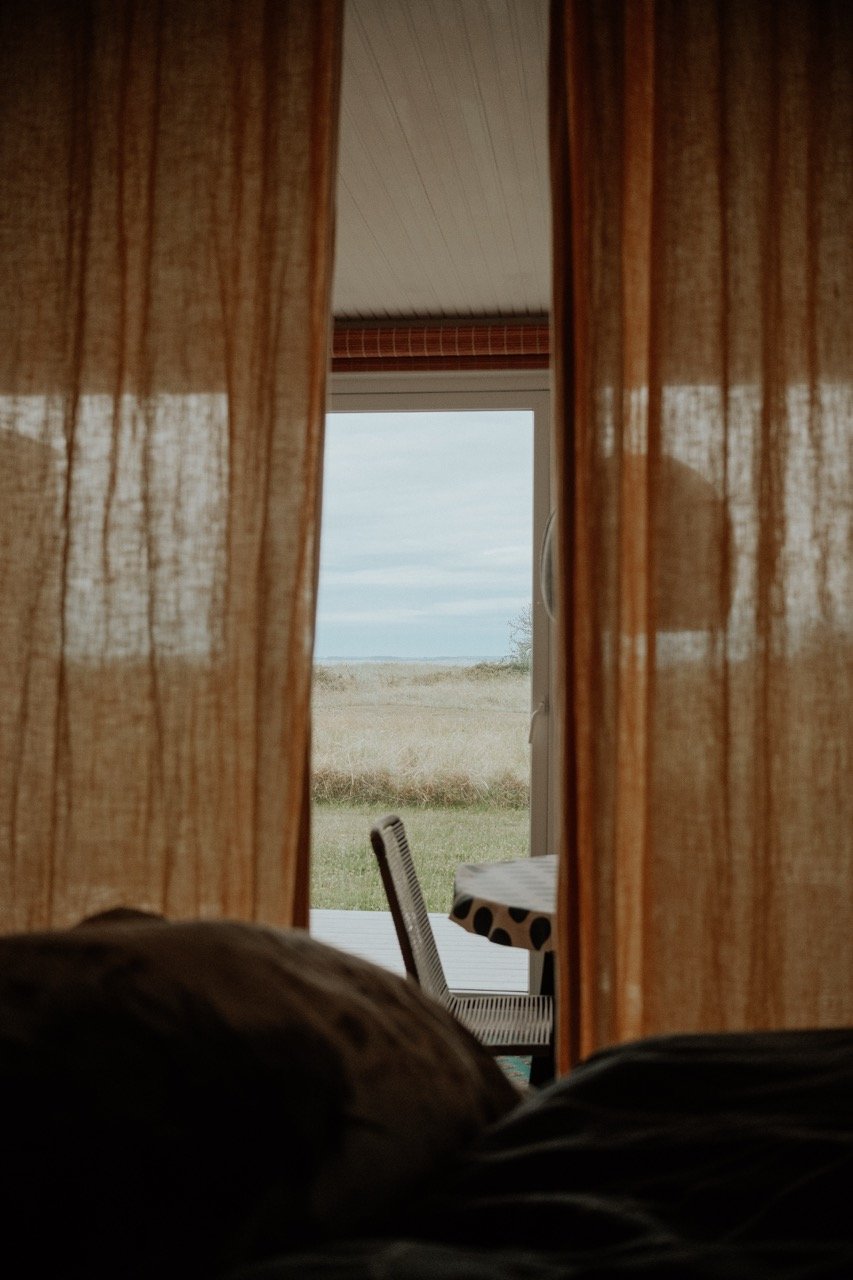 View through open curtains showing a chair with a polka dot tablecloth on a patio facing open field and sky.