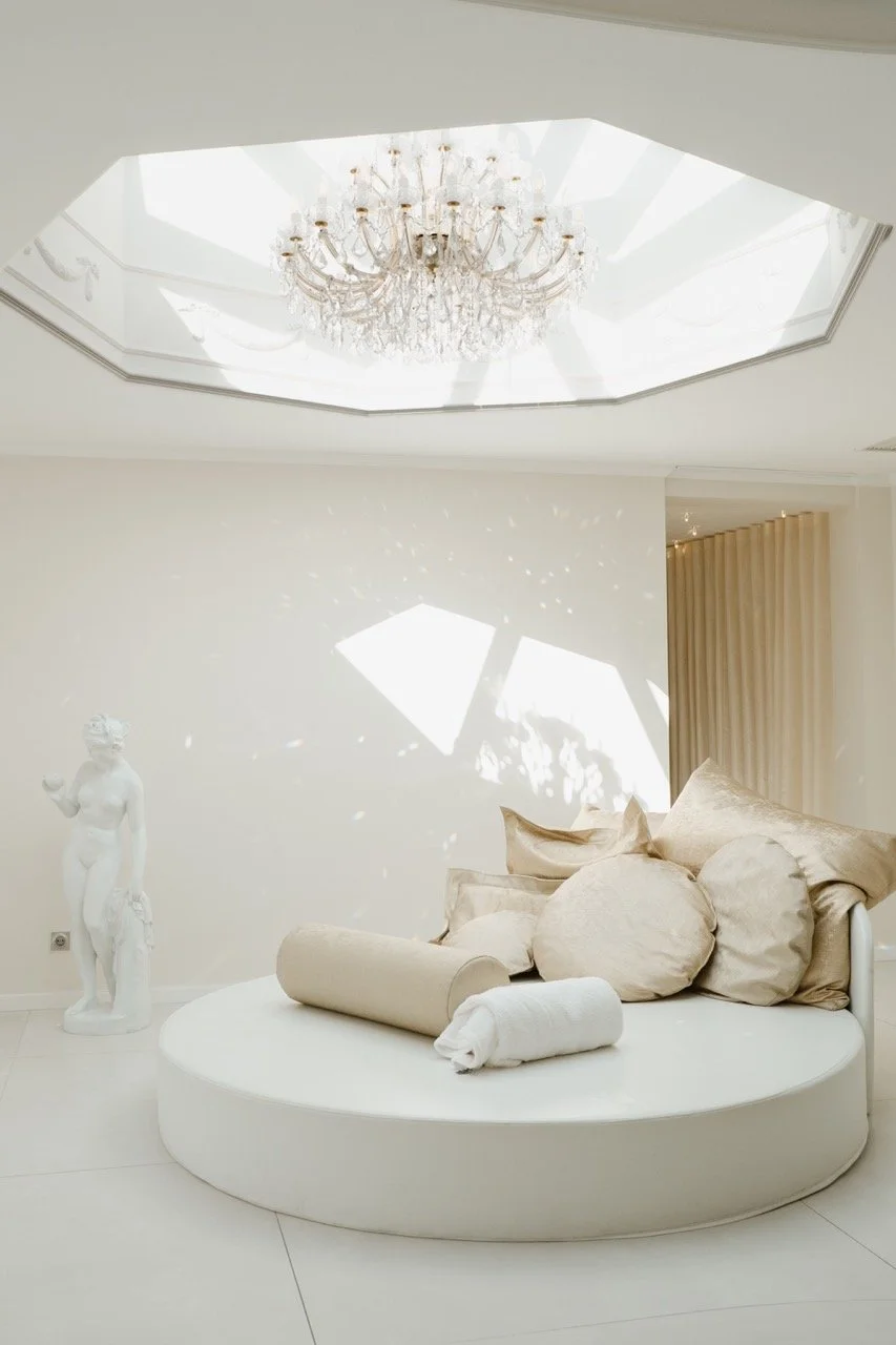 Elegant room with a white circular platform, beige pillows, and rolled-up towels, a statue of a woman, a crystal chandelier, and sunlight creating patterns on the wall.