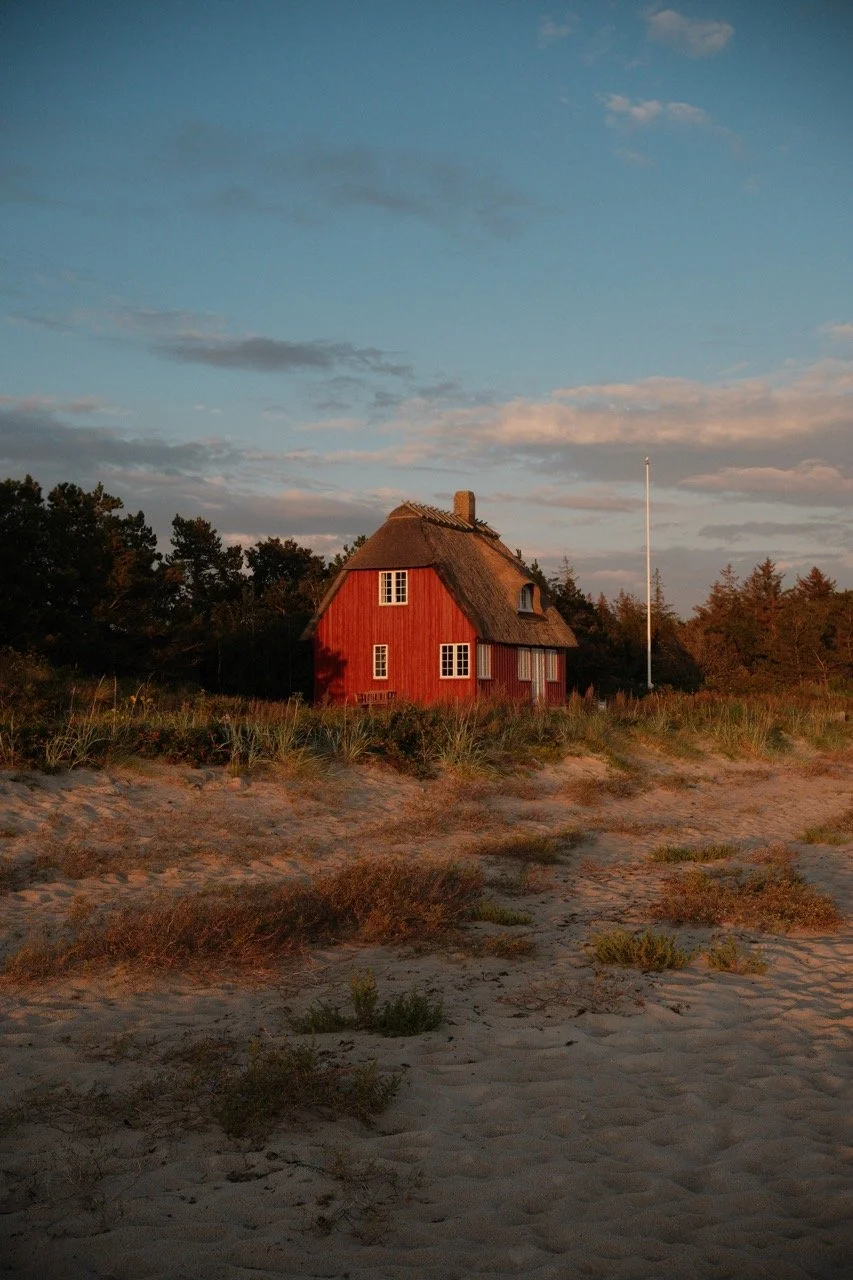 A red house with a thatched roof near a sandy beach, surrounded by bushes and trees, under a partly cloudy sky during sunset.