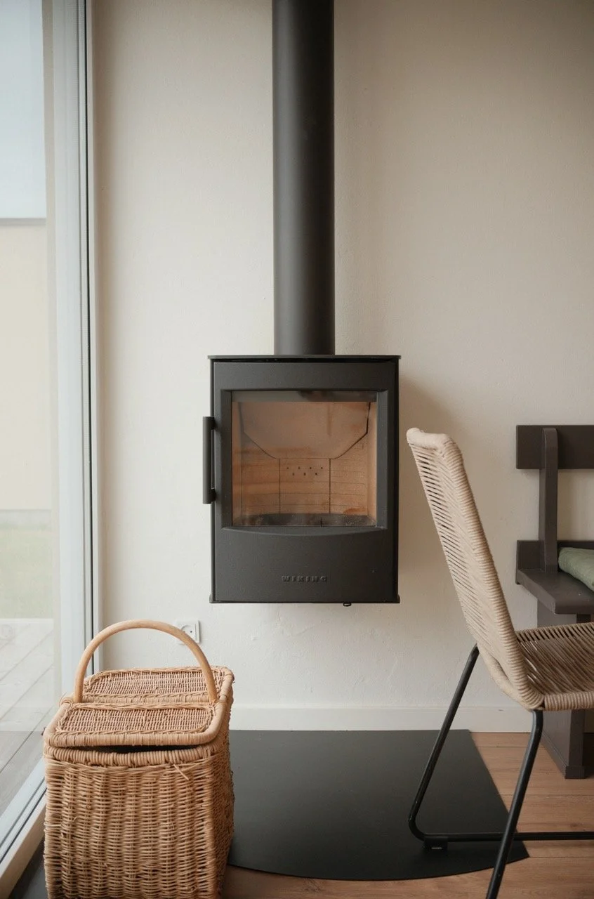 A modern black wood-burning stove with a glass door and a pipe extending upward, positioned against a white wall in a cozy indoor setting. To the left, a wicker basket with a handle, and to the right, a beige woven chair with a black frame.