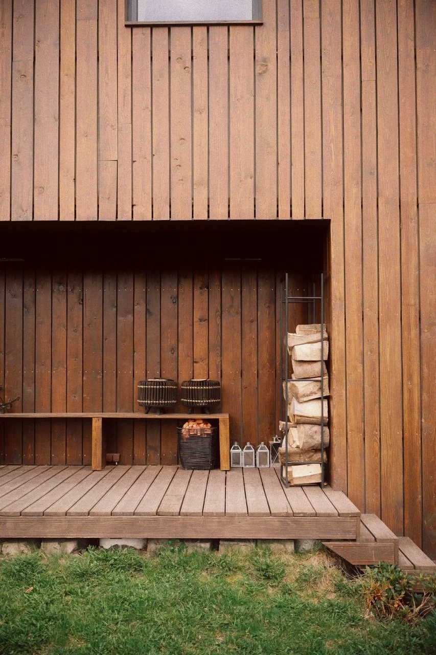Wooden deck in front of a wooden house wall with stacked firewood, lanterns, and decorative items.