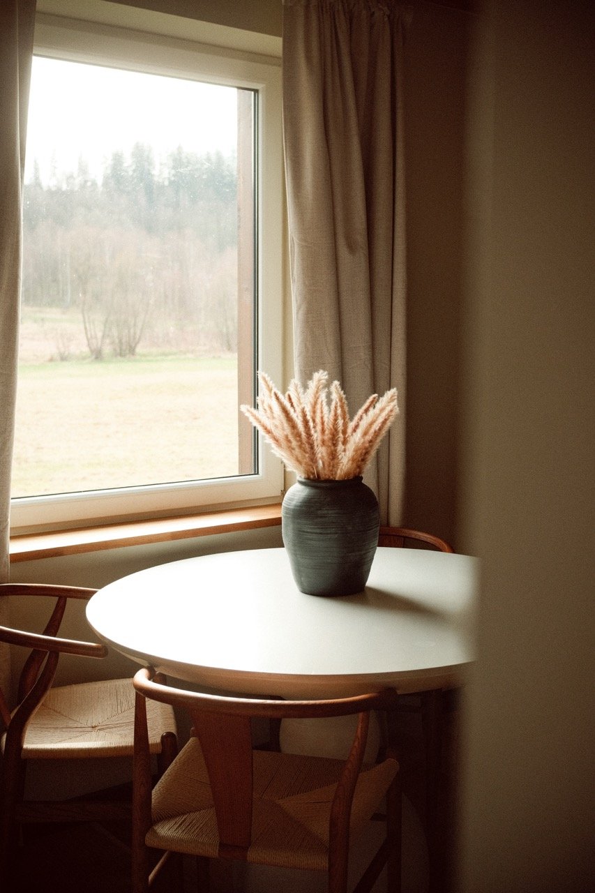 A round white dining table with a black vase containing pink pampas grass, placed near a window with beige curtains, and surrounded by wooden chairs with woven seats.