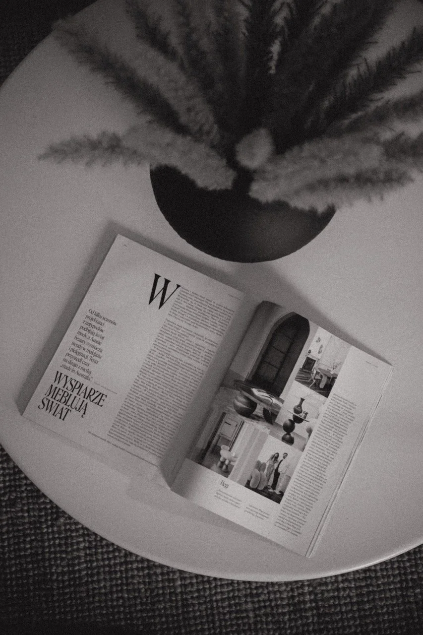 An open magazine on a white surface with a plant in a black pot in the background.