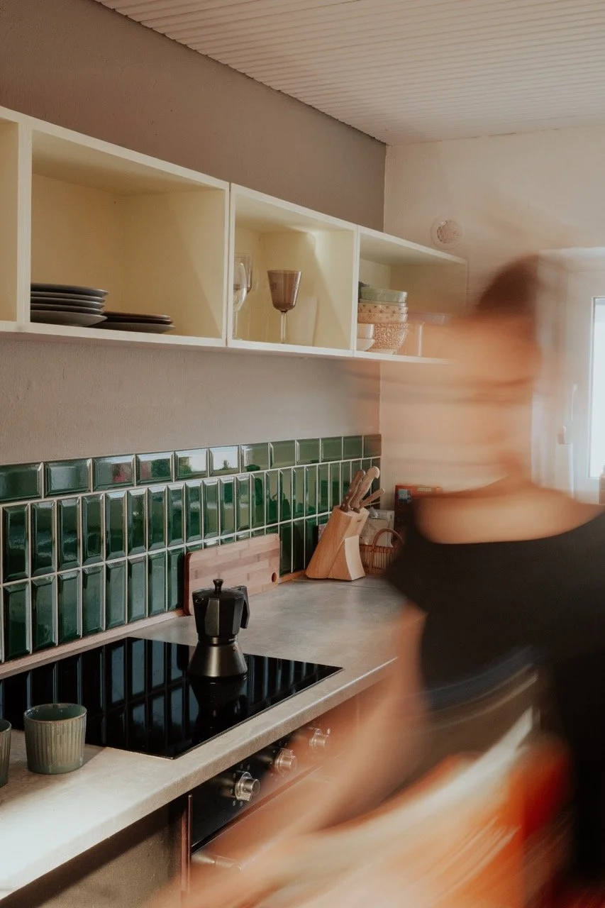 A person chopping something in a kitchen with green tiled backsplash, white open shelves, and a black stove with a moka pot on top.