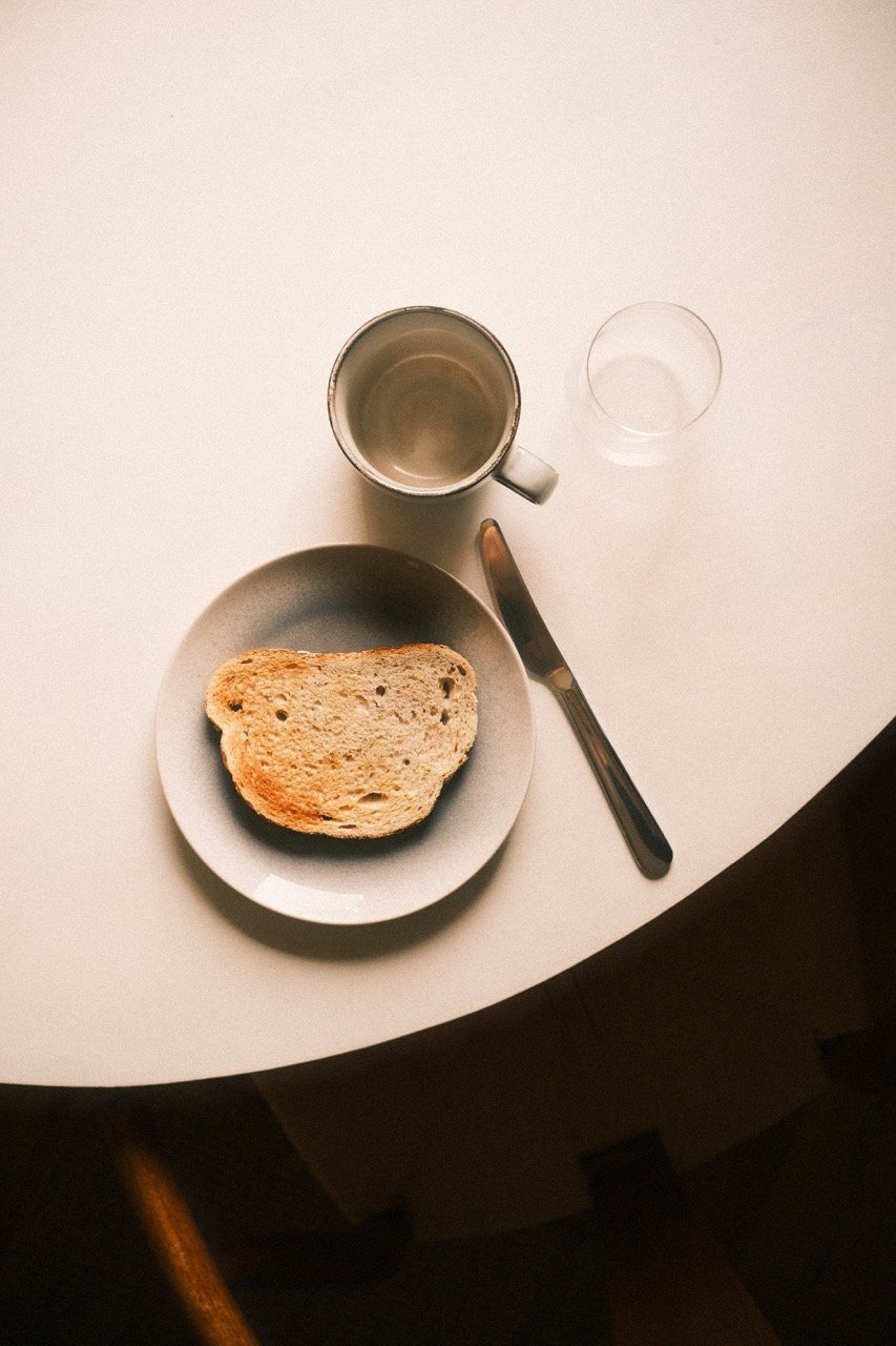 A meal on a white table, includes a slice of bread on a white plate, a knife, a metallic mug, and a glass of water.